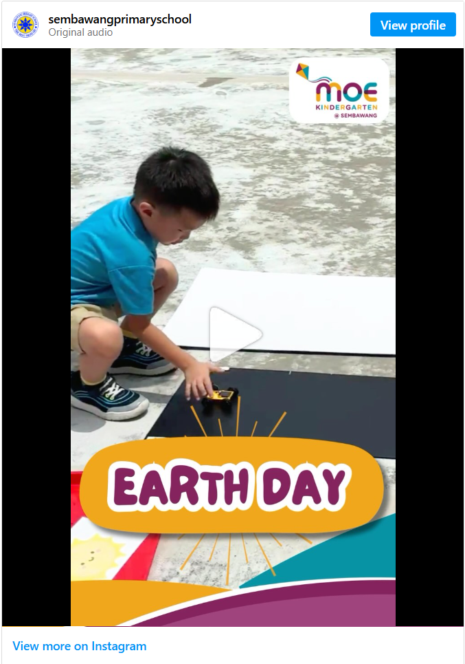 Instagram video post from sembawangprimaryschool (MOE Kindergarten @ Sembawang) titled "Earth Day," showing a boy in a blue uniform crouching on a textured outdoor surface, placing a small toy on a black mat with a sun-ray decoration and play button overlay indicating it's a video.