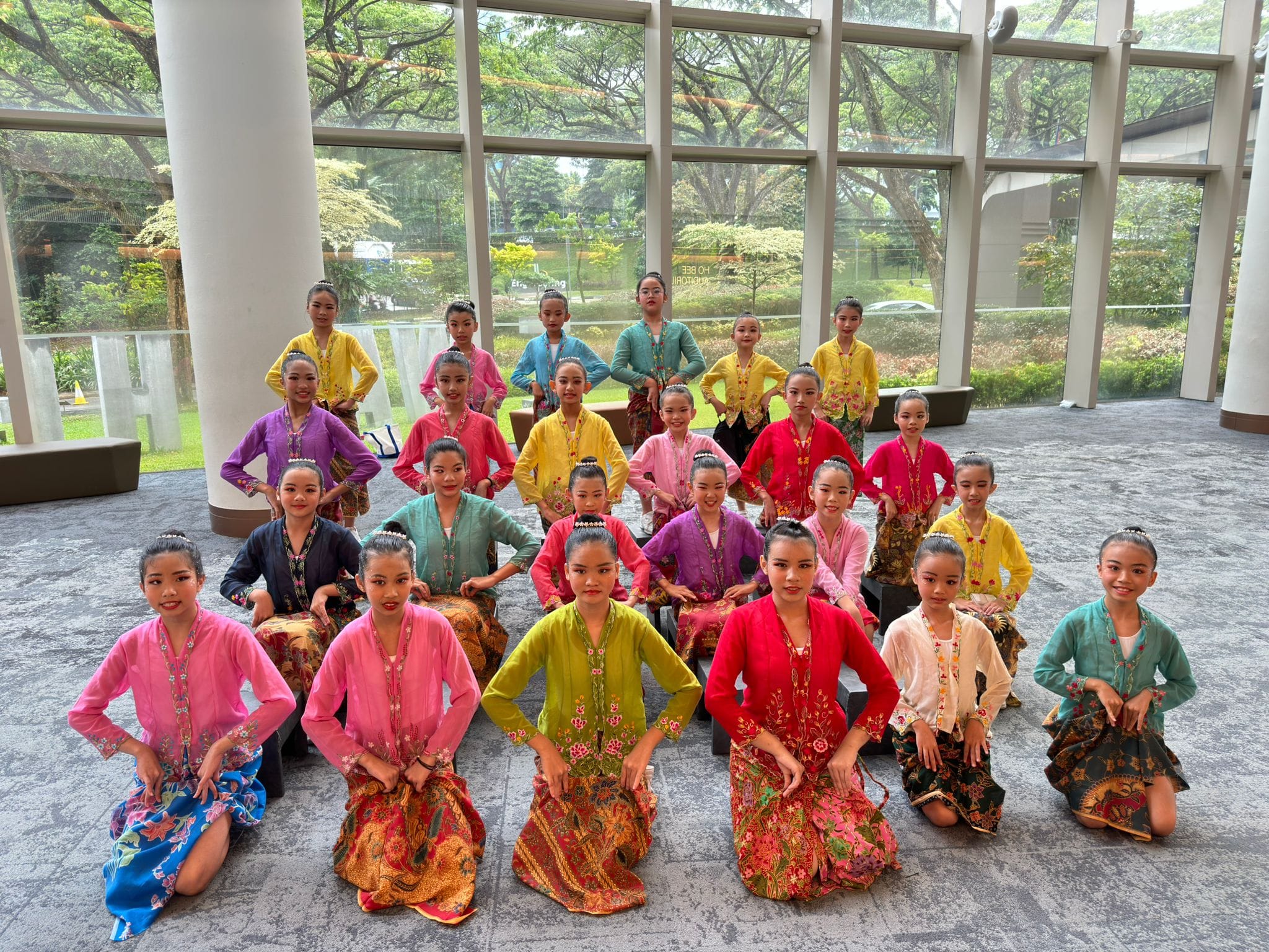 A group of young girls in colorful traditional Malay kebaya and batik skirts pose indoors in front of large windows.