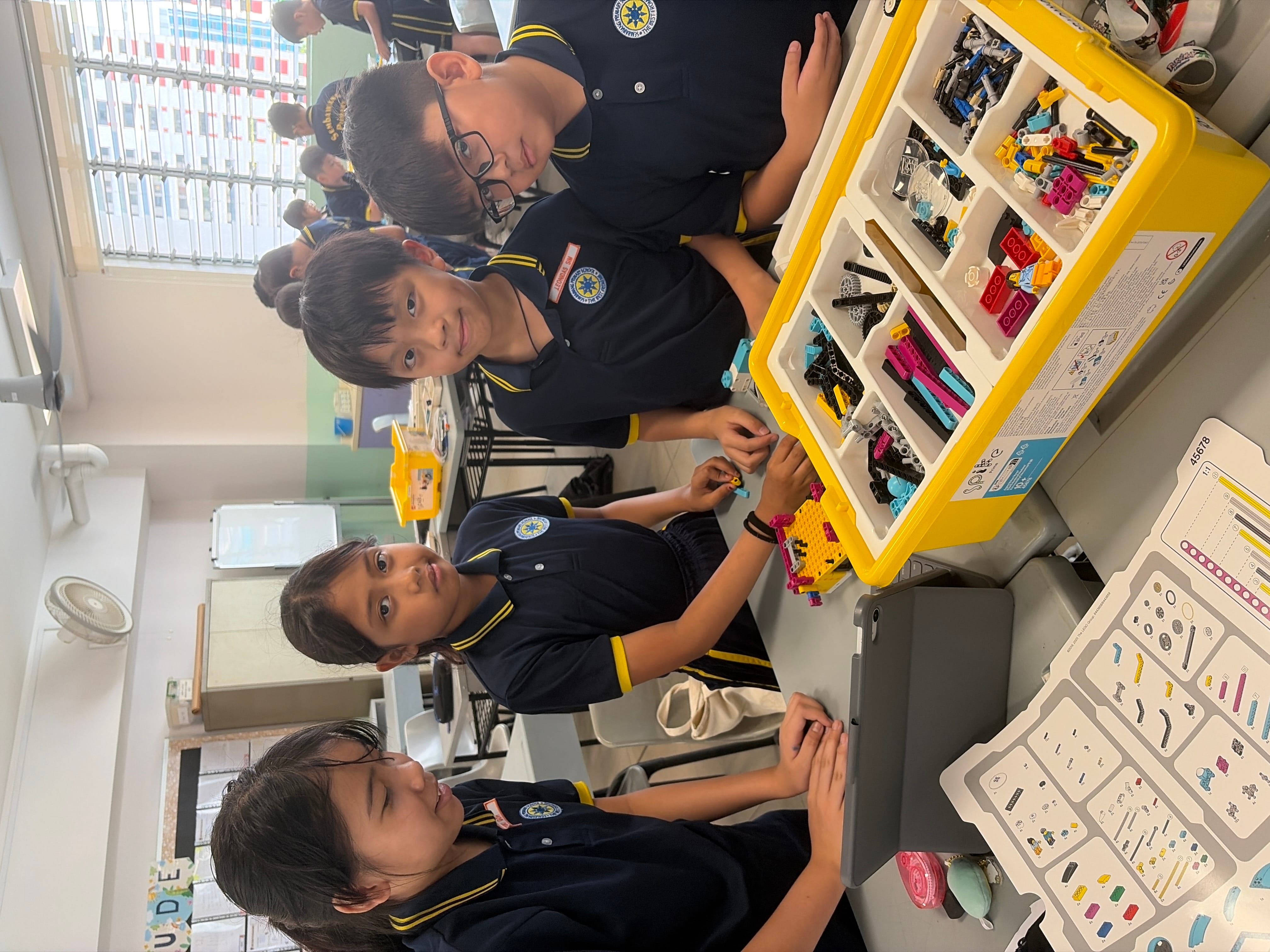Four school children in navy uniforms work together around a yellow bin of colorful LEGO bricks in a classroom.