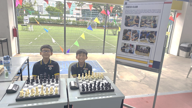 Two boys sit smiling behind chessboards set up on tables at an outdoor booth, with a "Chess Club" information poster displayed beside them and a school field visible in the background, decorated with colourful bunting flags.