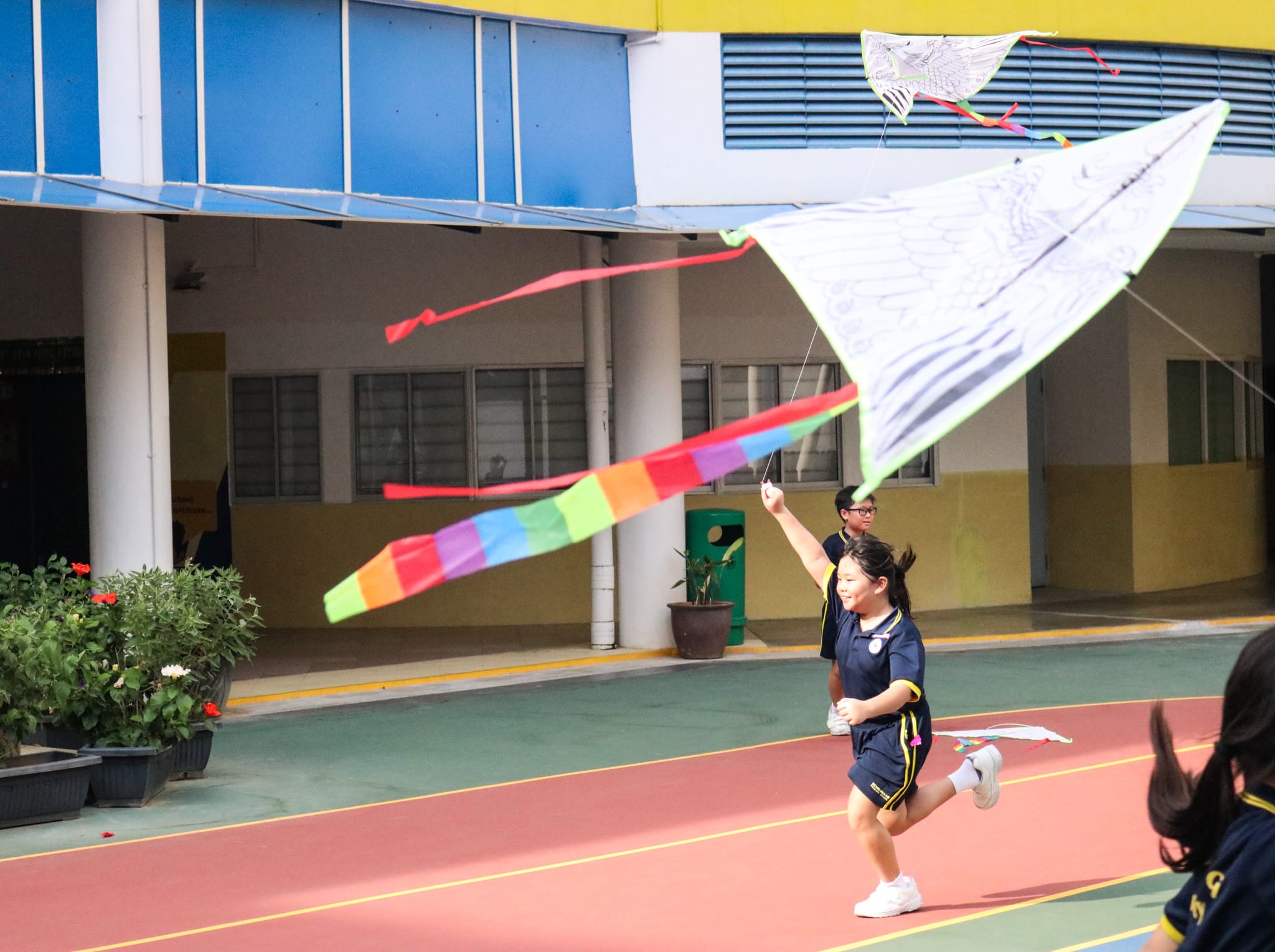 A girl in a navy uniform runs across a school courtyard, flying a large white kite with colorful tails.