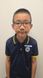 A young boy in glasses wearing a navy school uniform with yellow trim and a name tag smiles at the camera against a plain background.