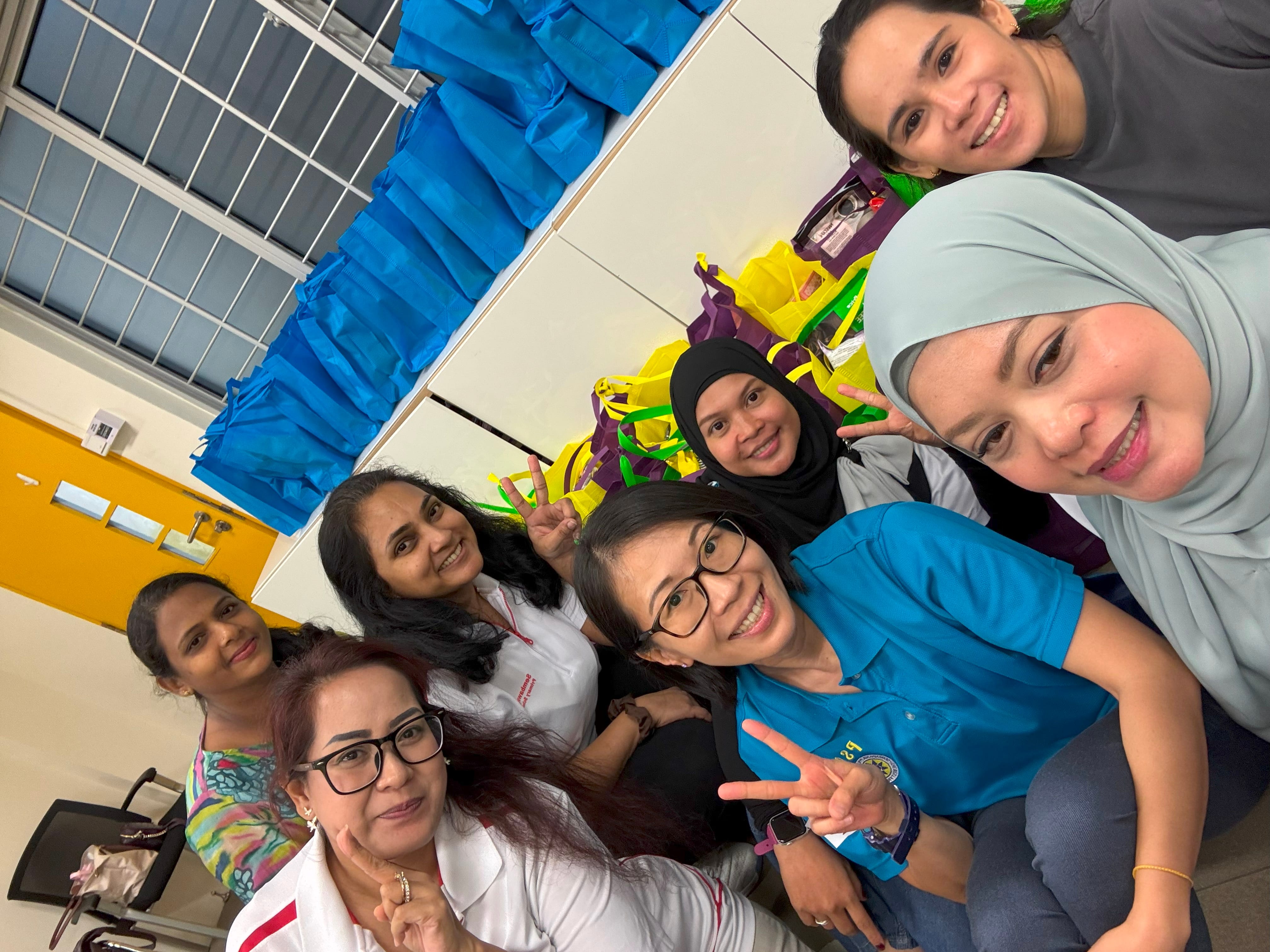 Six smiling women pose for a selfie indoors near blue and yellow reusable bags, some making peace signs.