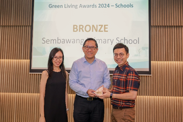 Two men and a woman posing on stage, with one man receiving an award trophy, in front of a screen displaying "Green Living Awards 2024 – Schools, BRONZE, Sembawang Primary School."
