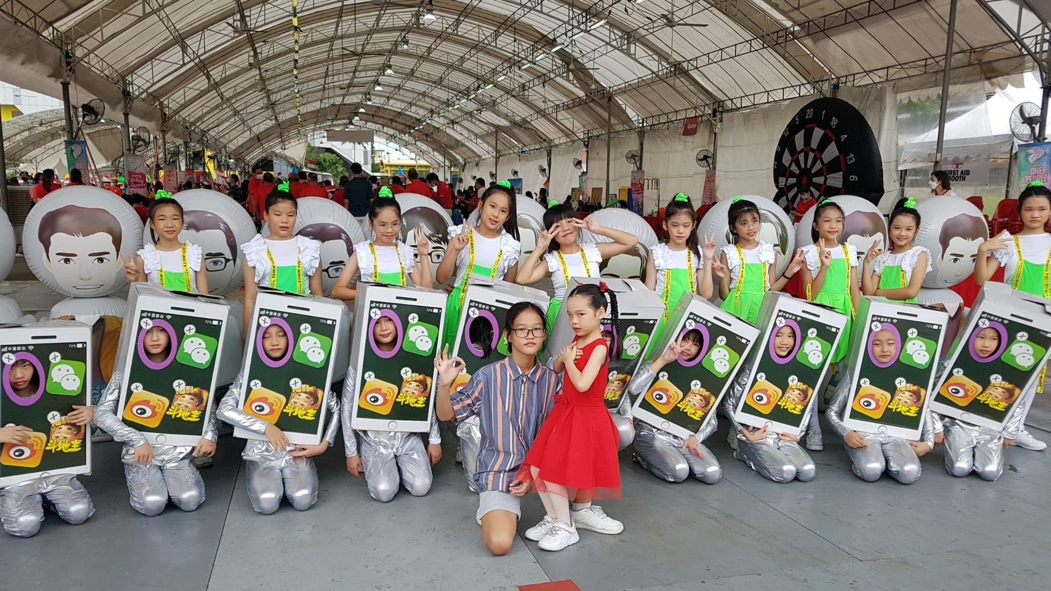 Children in costumes shaped like smartphones pose together at an indoor event under a large tent.