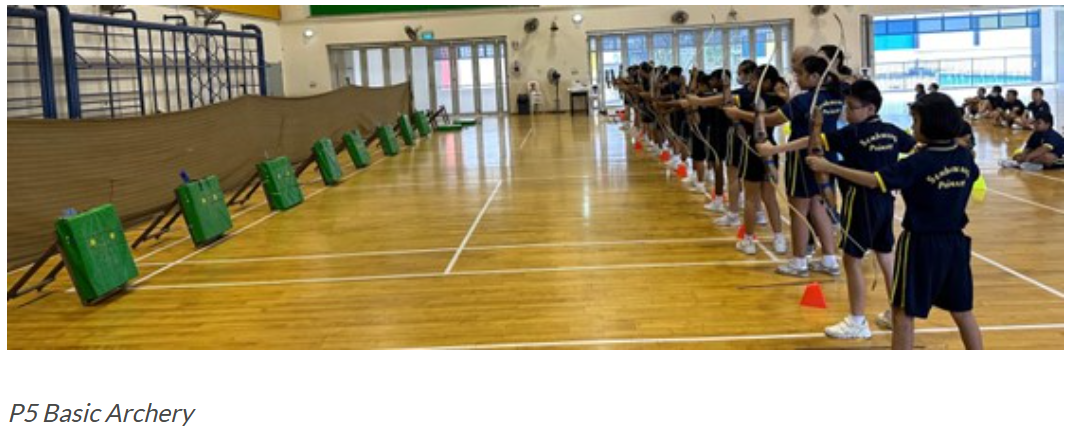 P5 Basic Archery: A line of P5 students in school PE attire practice archery in the school hall, aiming bows at green target boxes set up along one wall.