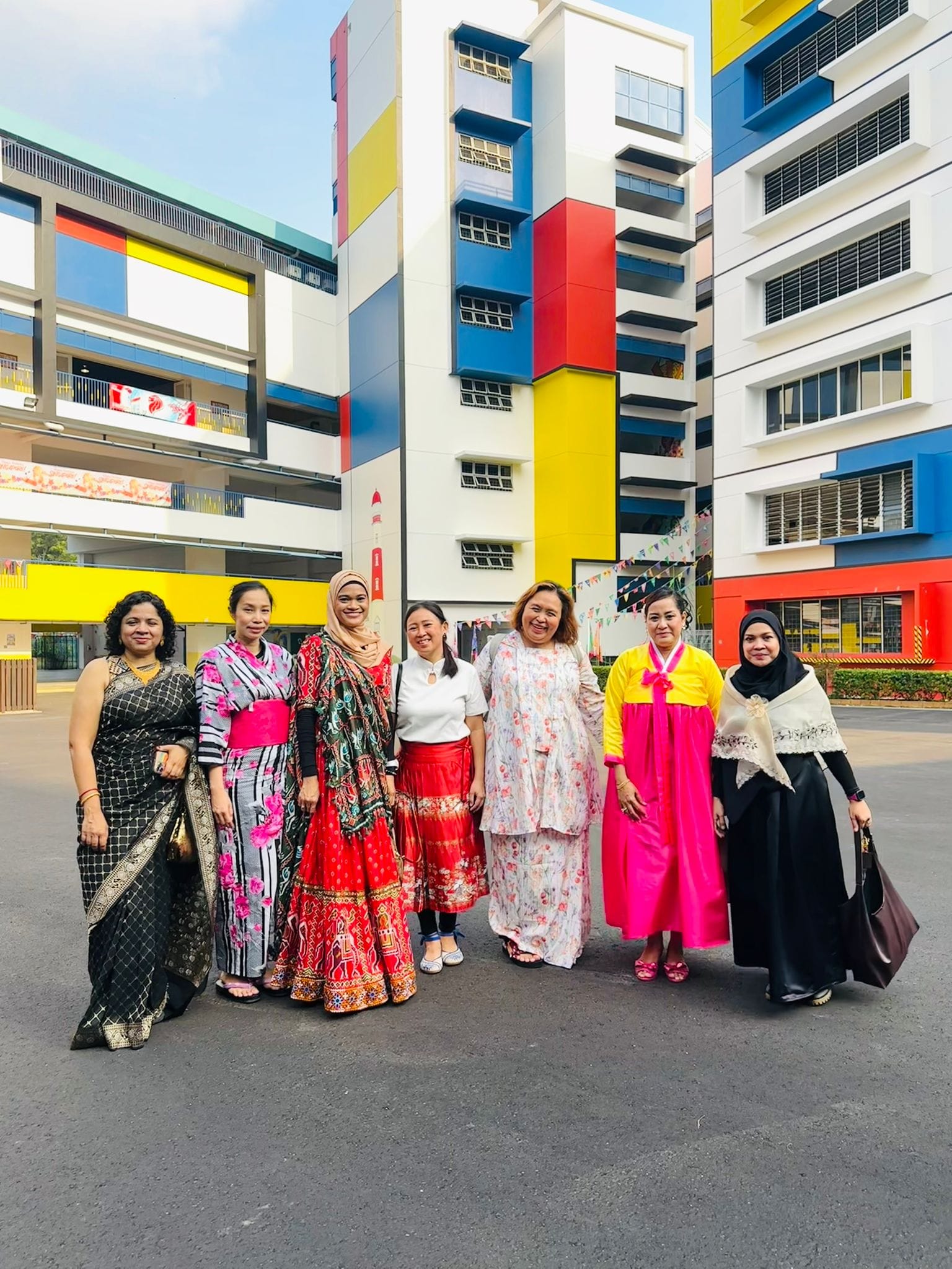 Seven women in colorful traditional cultural outfits stand together in front of a bright multi-colored building.