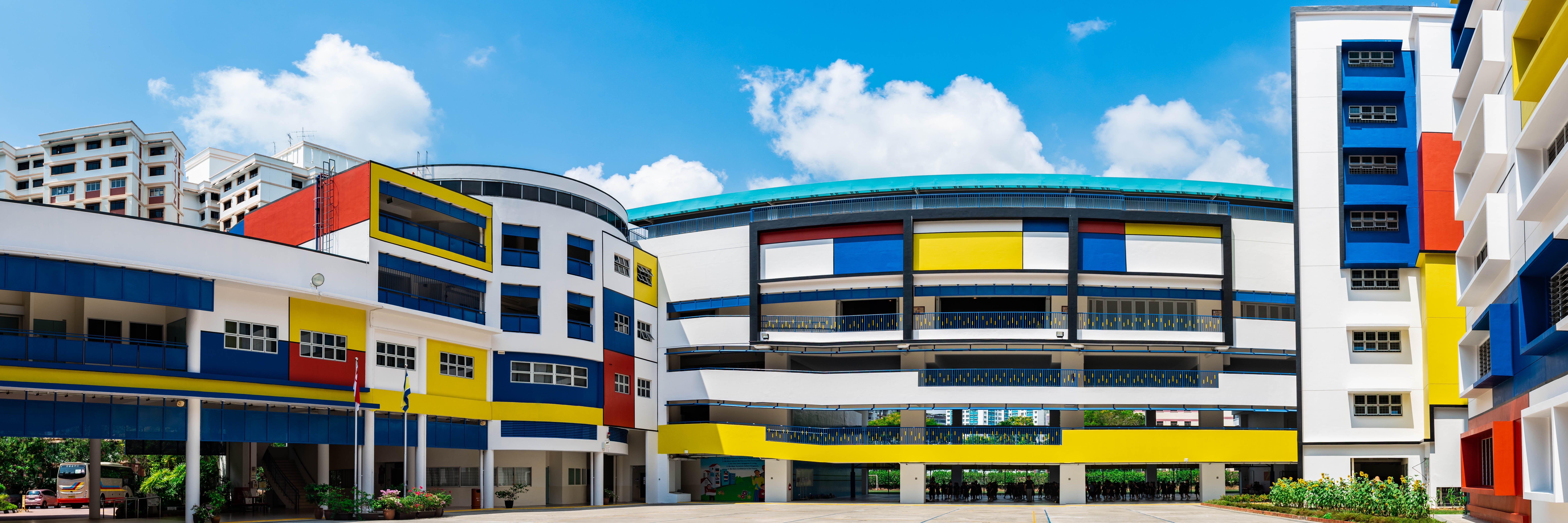 Colorful school buildings with red, yellow, and blue panels under a bright blue sky with clouds.