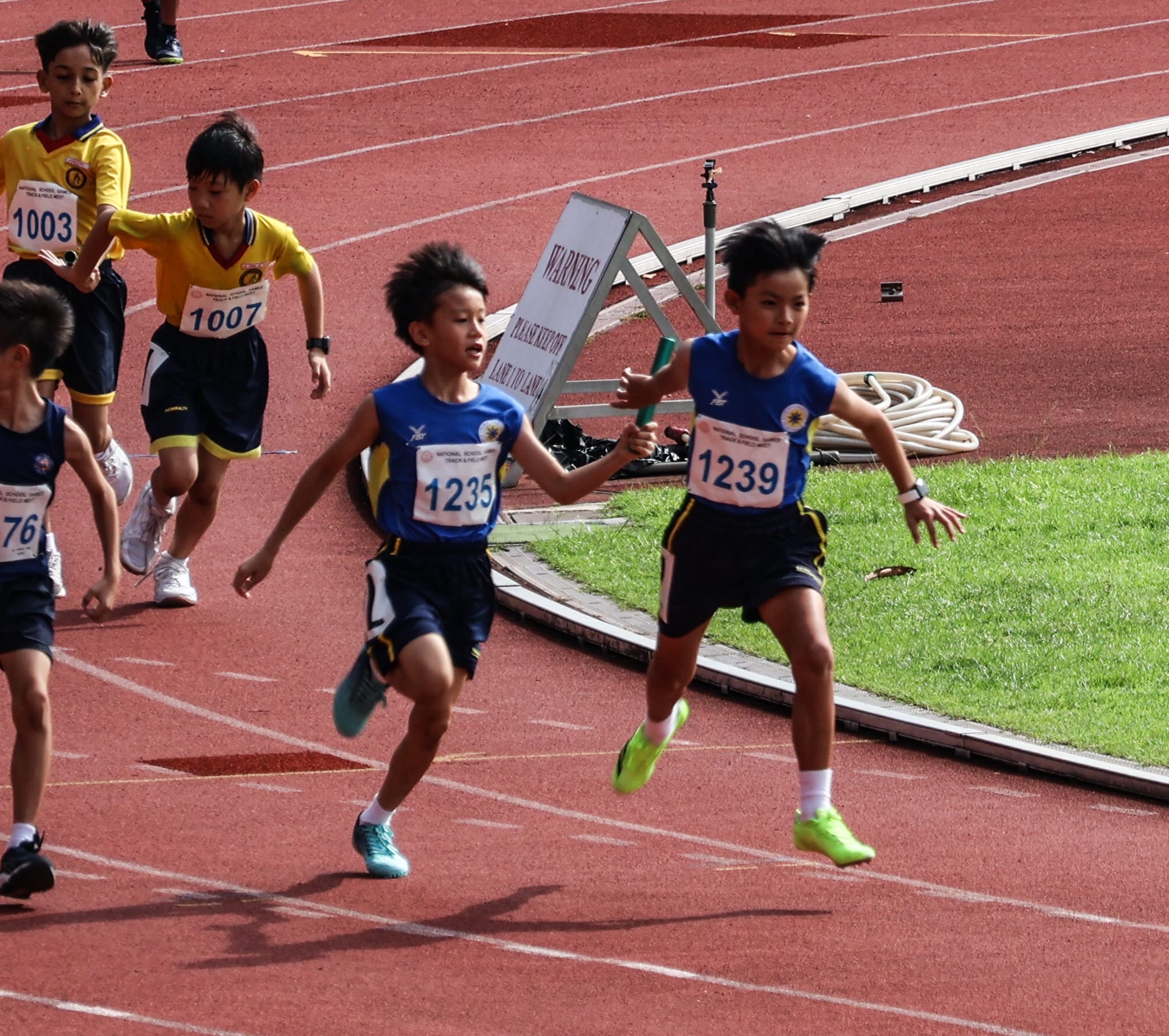 A relay runner in blue passing the baton to a teammate mid-stride, with other teams in yellow and navy also mid-exchange in the background.