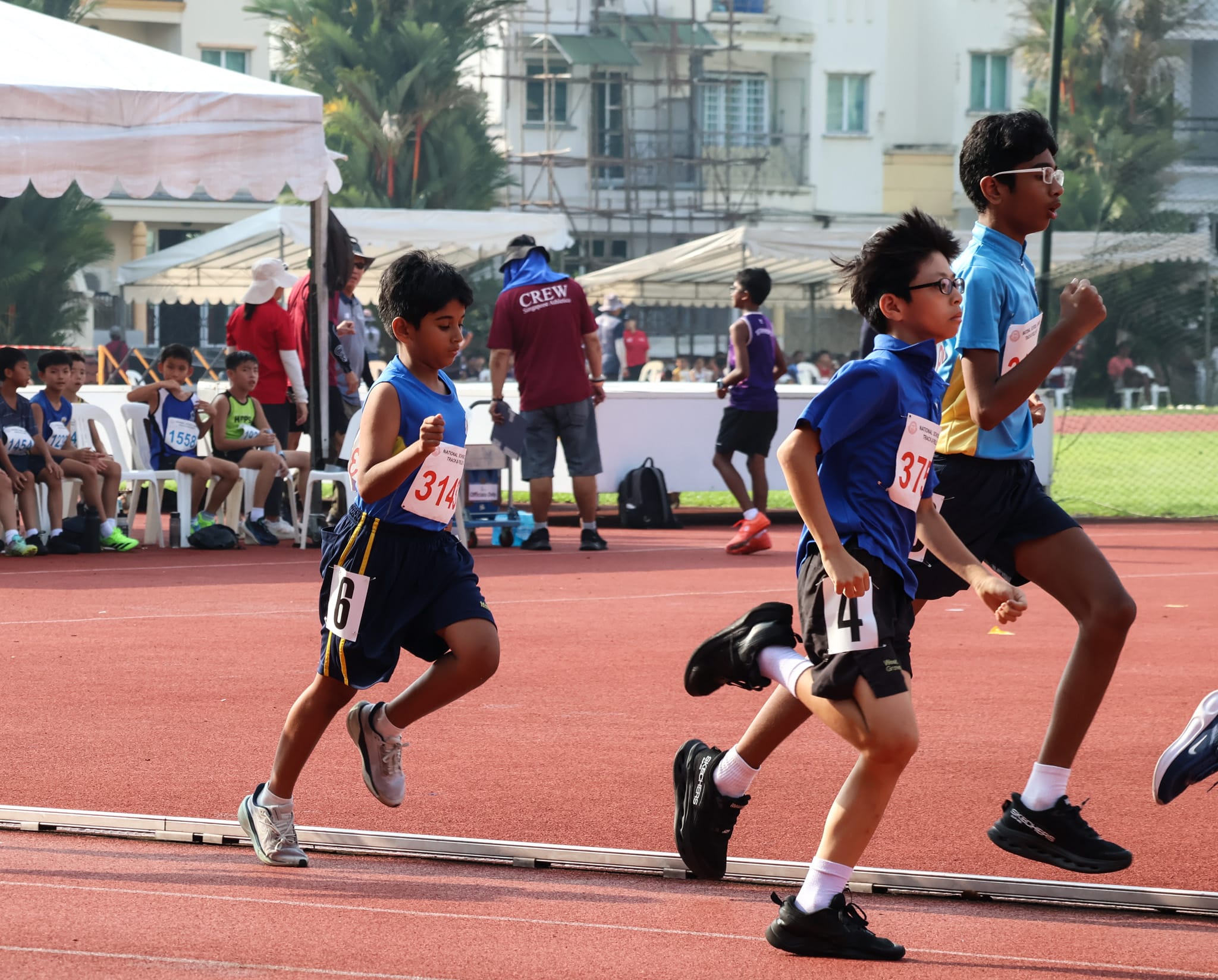 A group of runners mid-stride during a race at the National School Games Track and Field Meet, with officials and spectators seated along the track in the background.