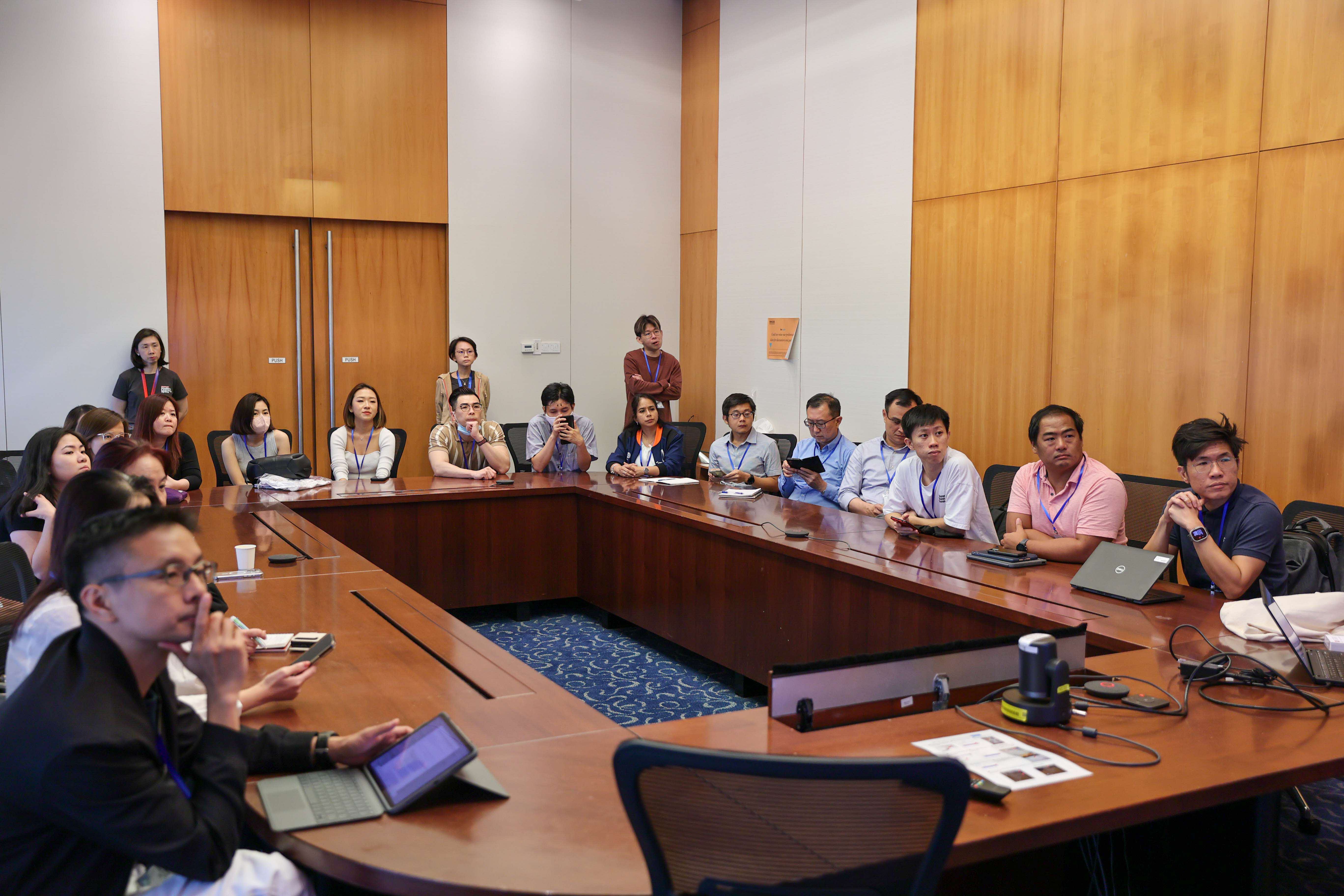 A large group of people sit and stand around a wooden conference table with laptops and phones during a meeting.