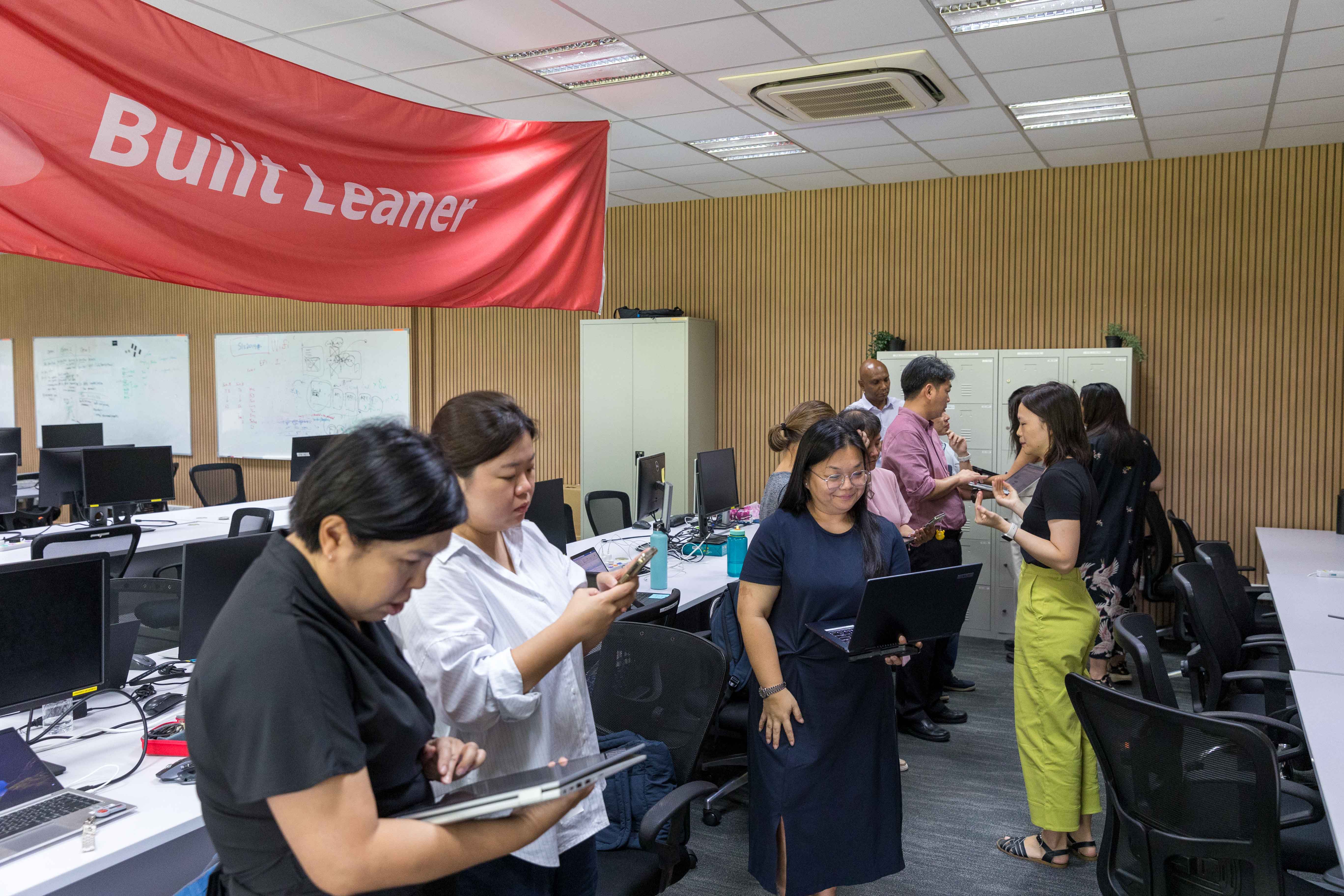 Group of people standing in an office room with a red "Built Leaner" banner, using laptops and phones.