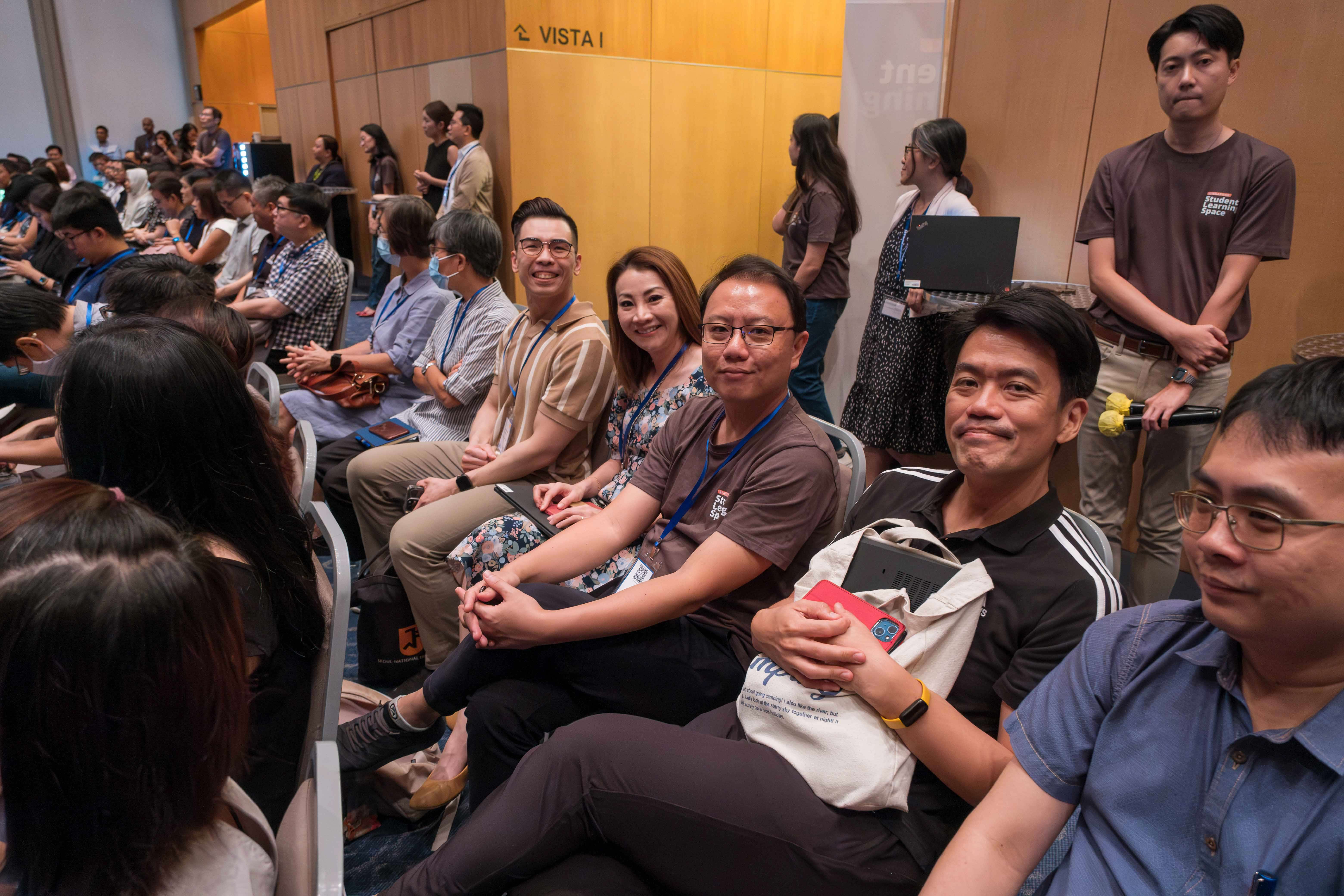 A large group of people seated and standing in a conference room labeled Vista I, smiling at the camera.