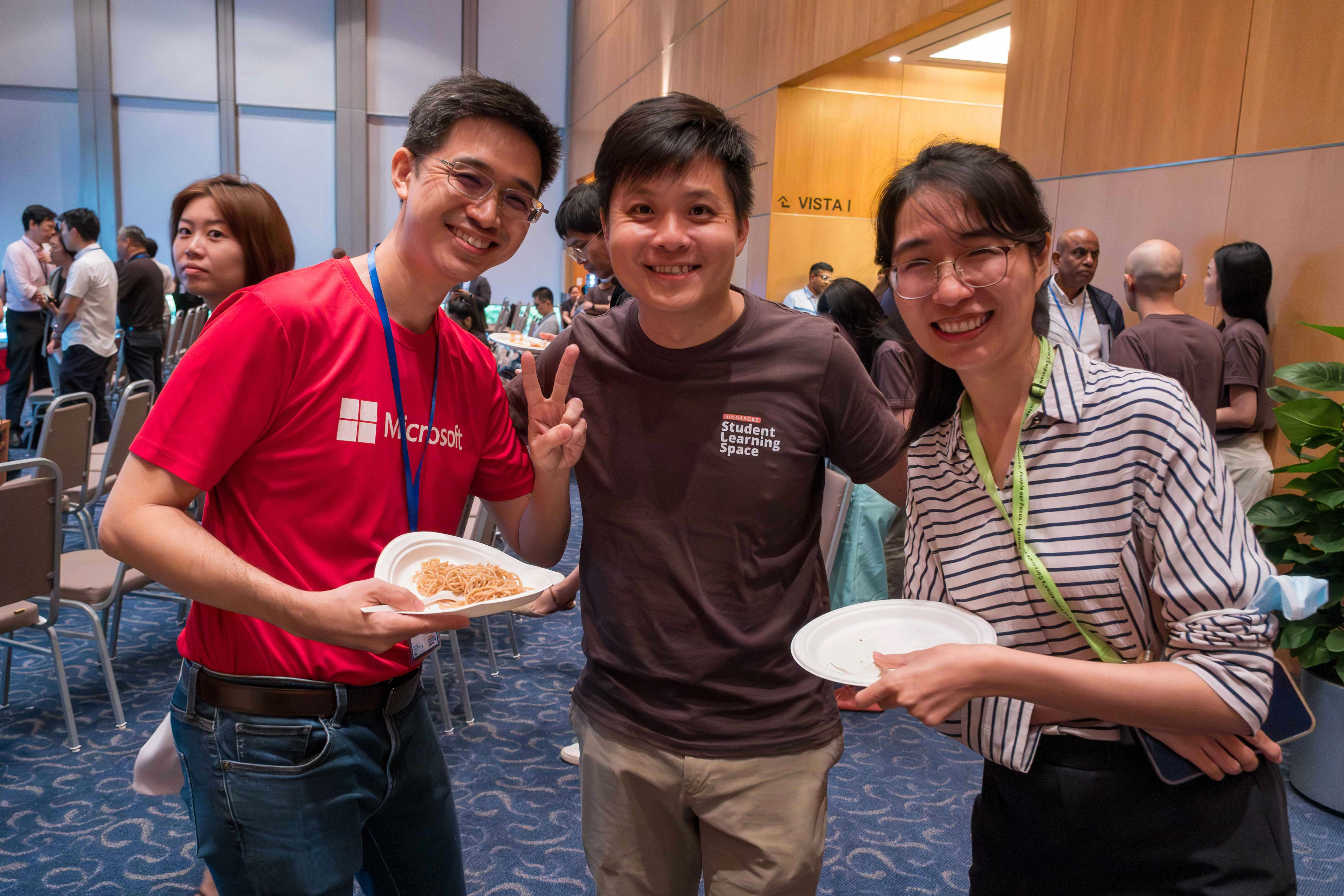 Three smiling people pose at an indoor event, two holding food plates, one in a red Microsoft shirt.