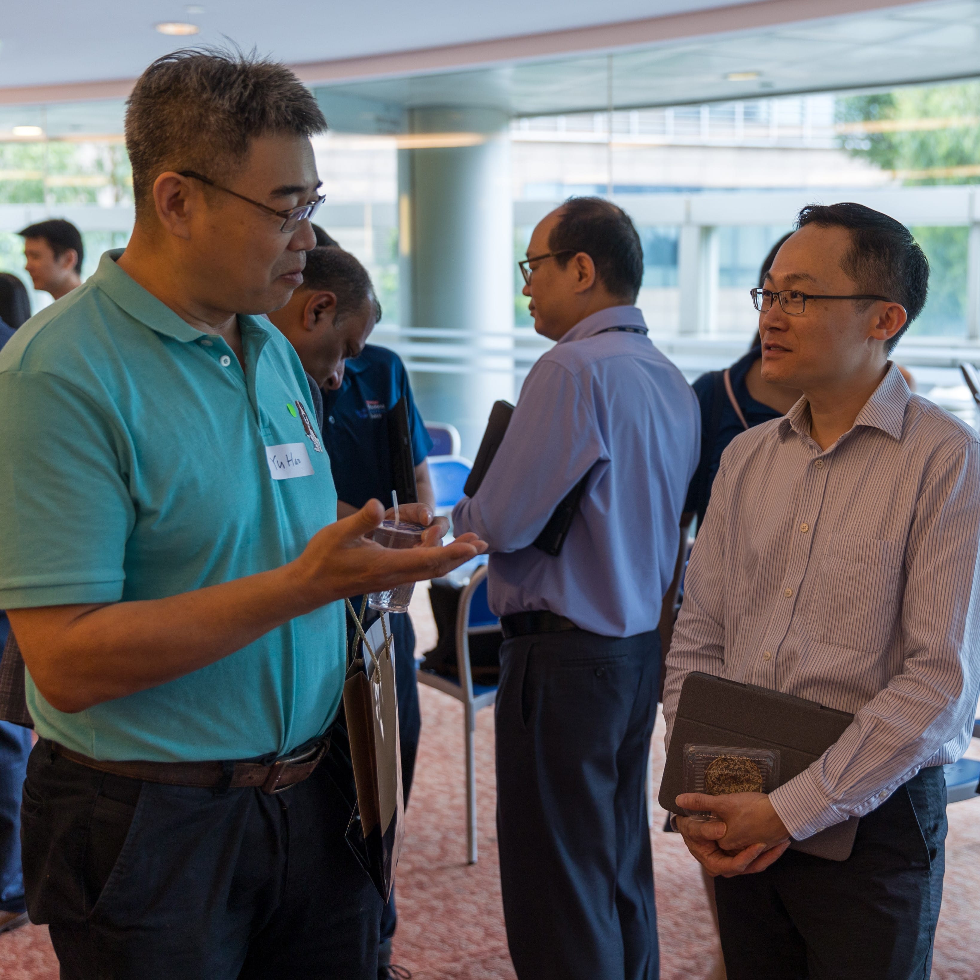 Two men with glasses converse at an indoor event, with other attendees visible in the background.