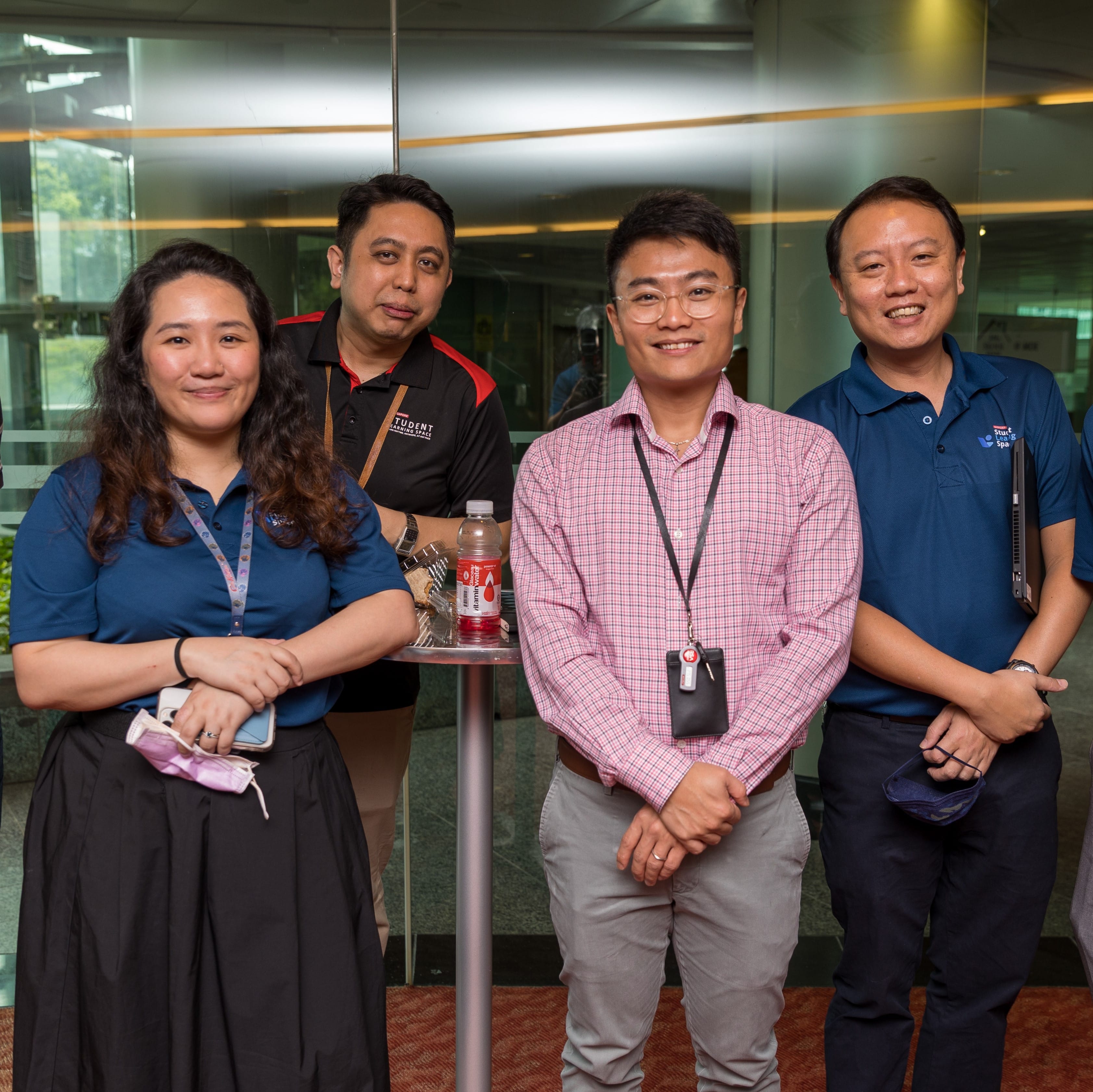 Four adults smiling outdoors near a glass building entrance, one woman in blue polo and three men wearing lanyards.