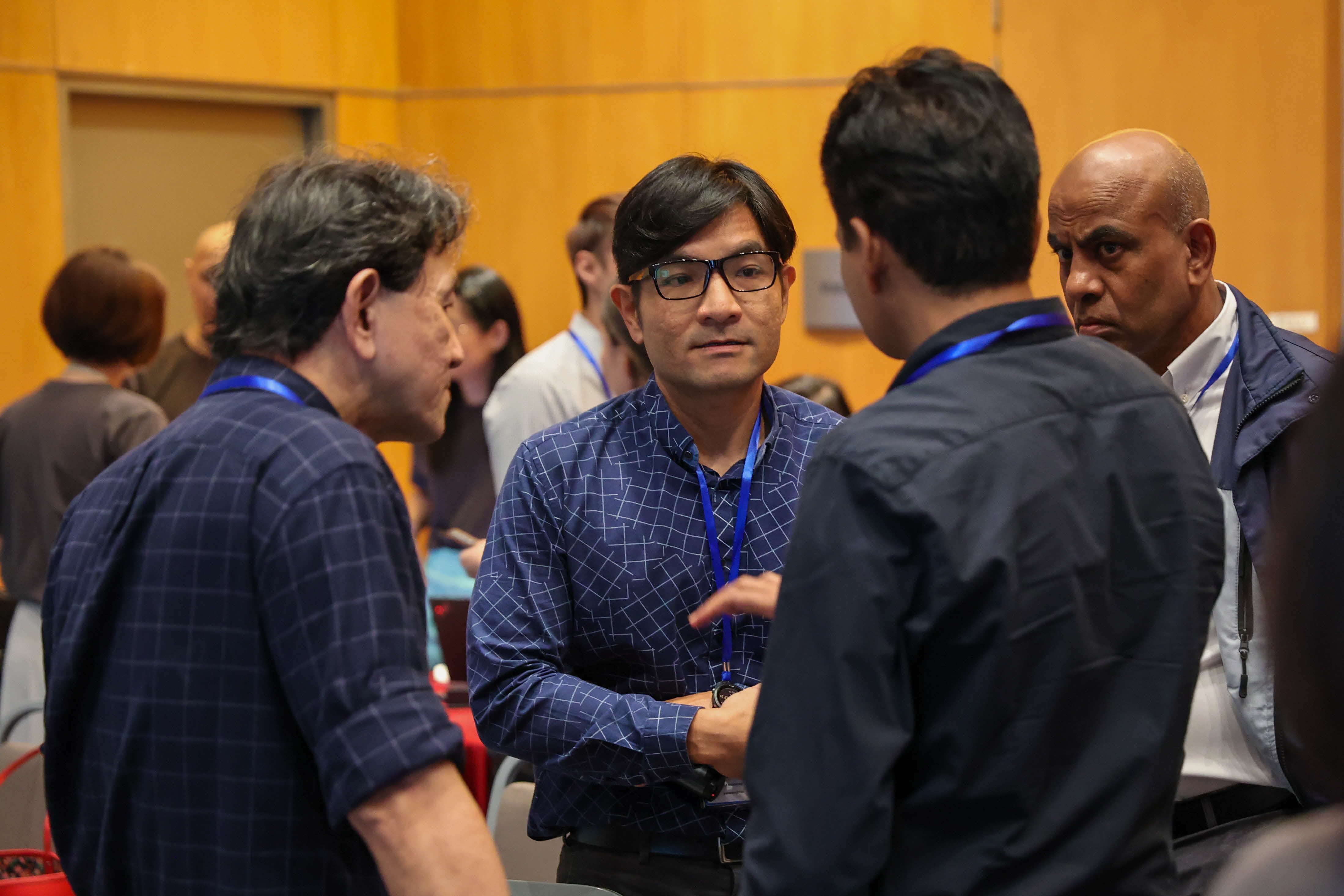 Four men wearing blue lanyards stand in a group conversation at a conference event inside a wood-paneled room.