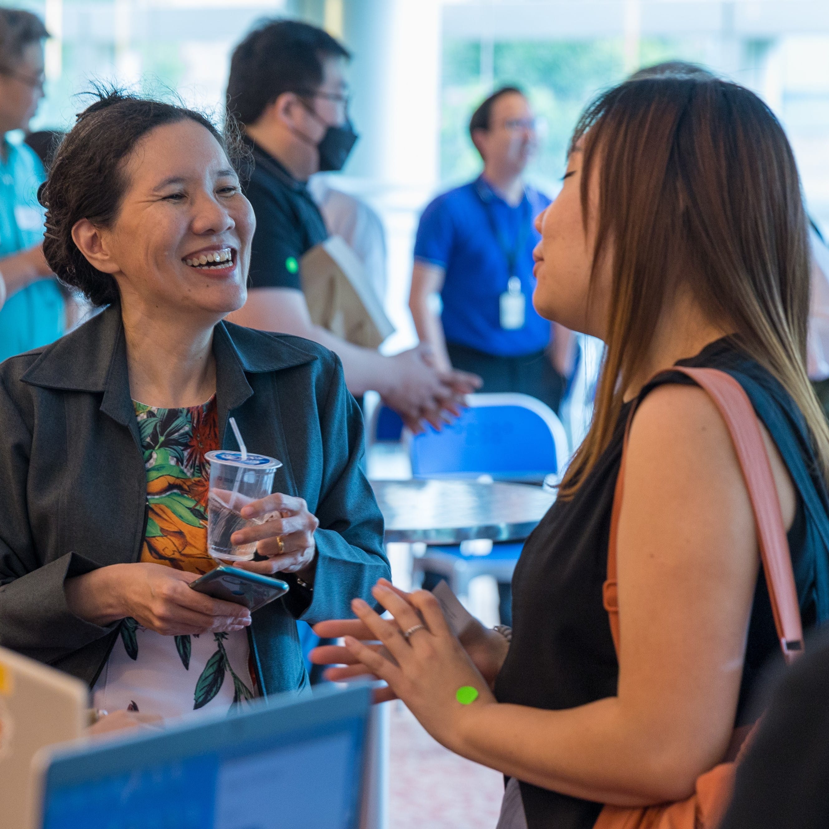Two women chat and laugh at an indoor event, one holding a plastic drink cup, with several people visible in the background.