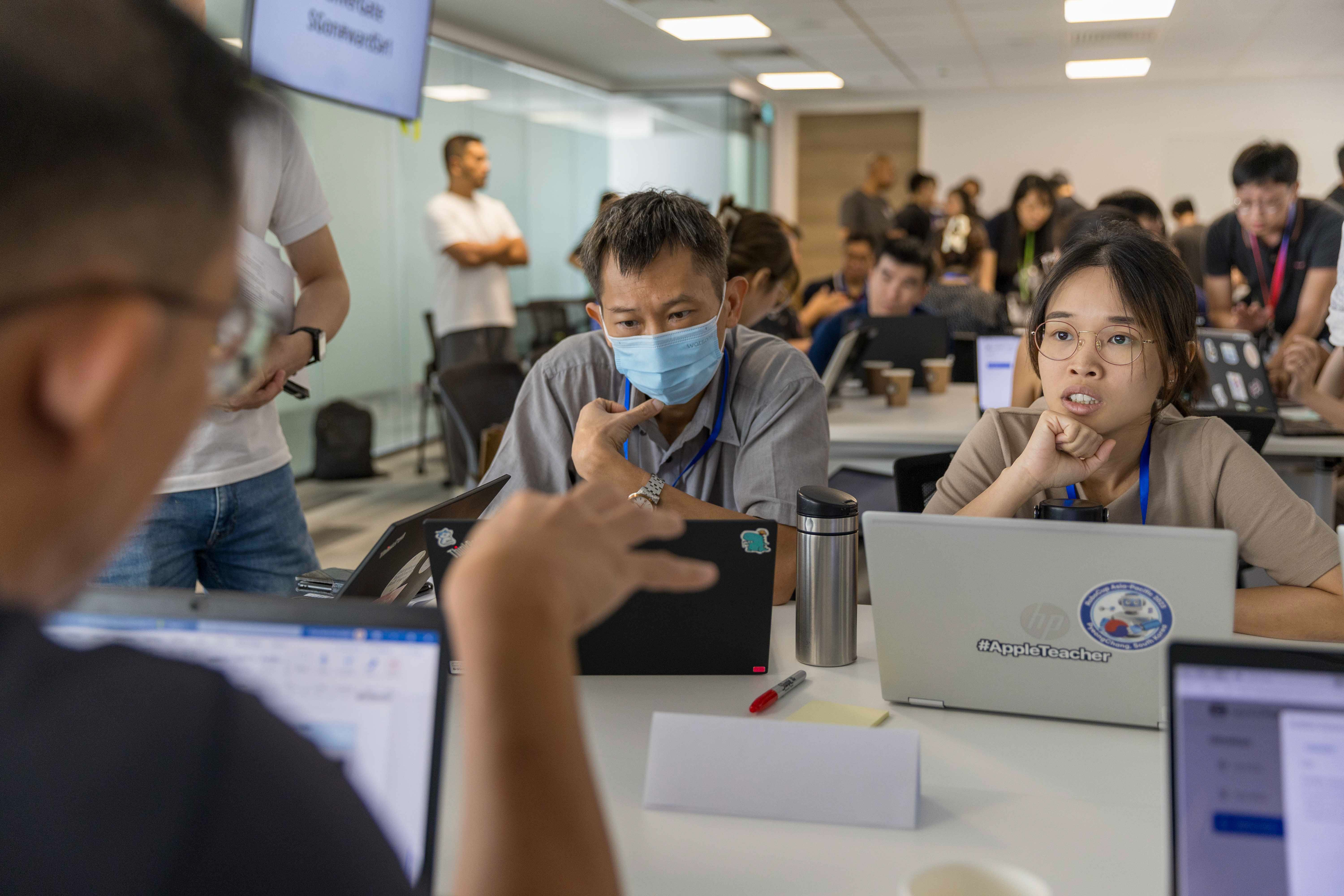 People sitting at tables with laptops in a busy workshop, one man wearing a blue face mask, listening attentively.