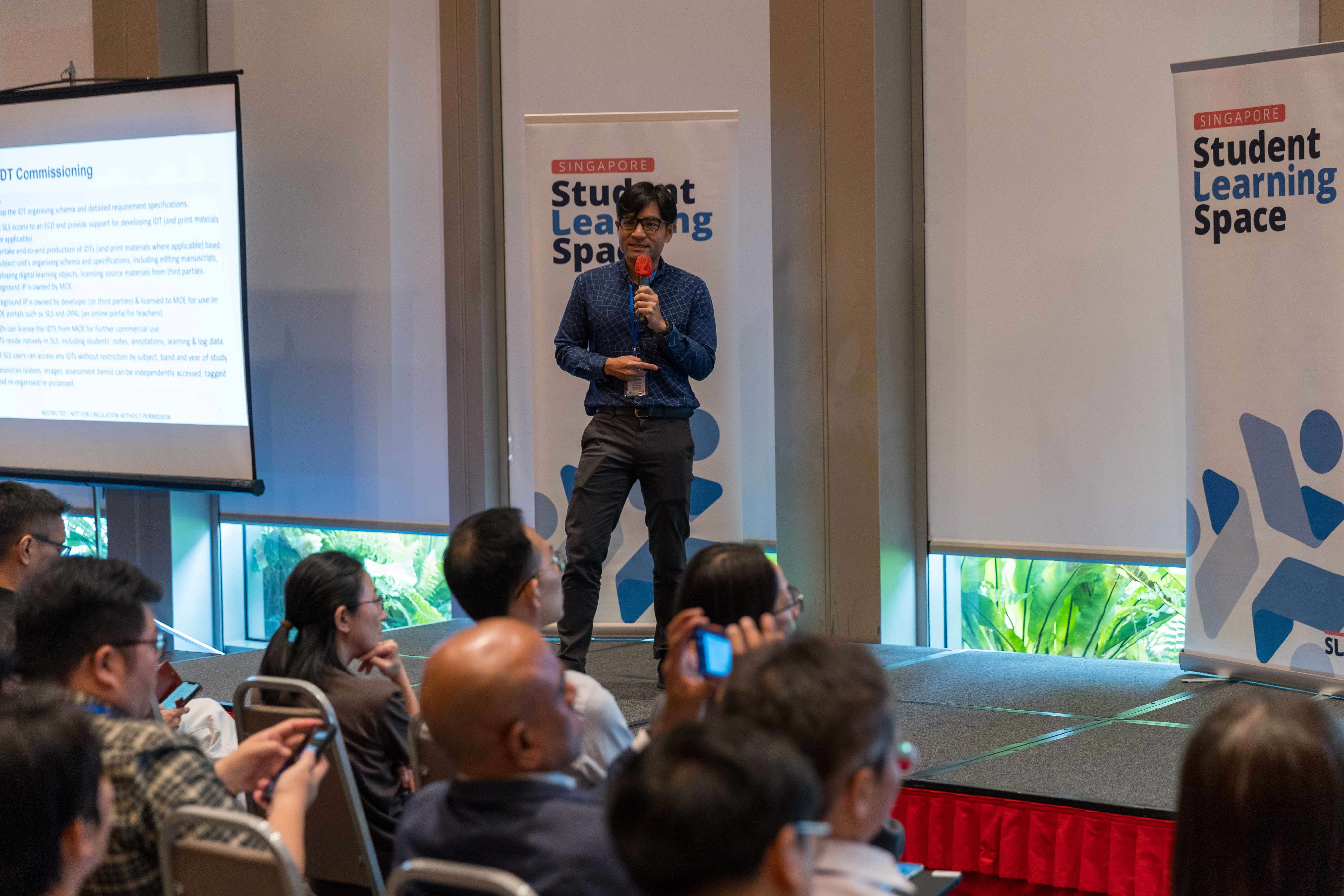 A man holding a microphone speaks on stage at a Singapore Student Learning Space event before a seated audience.
