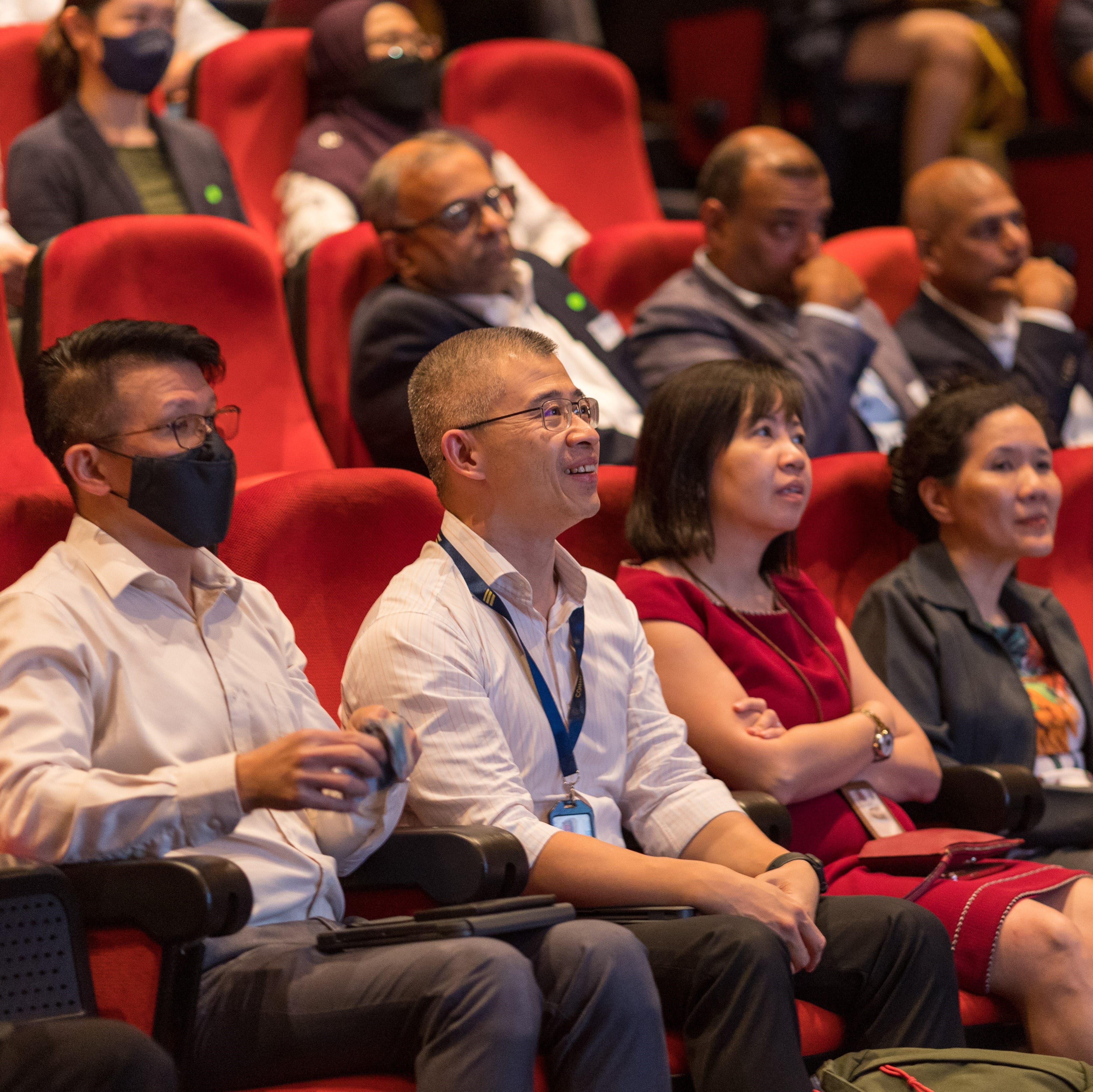 Audience members seated in red chairs at an event, some wearing masks, looking attentively forward.
