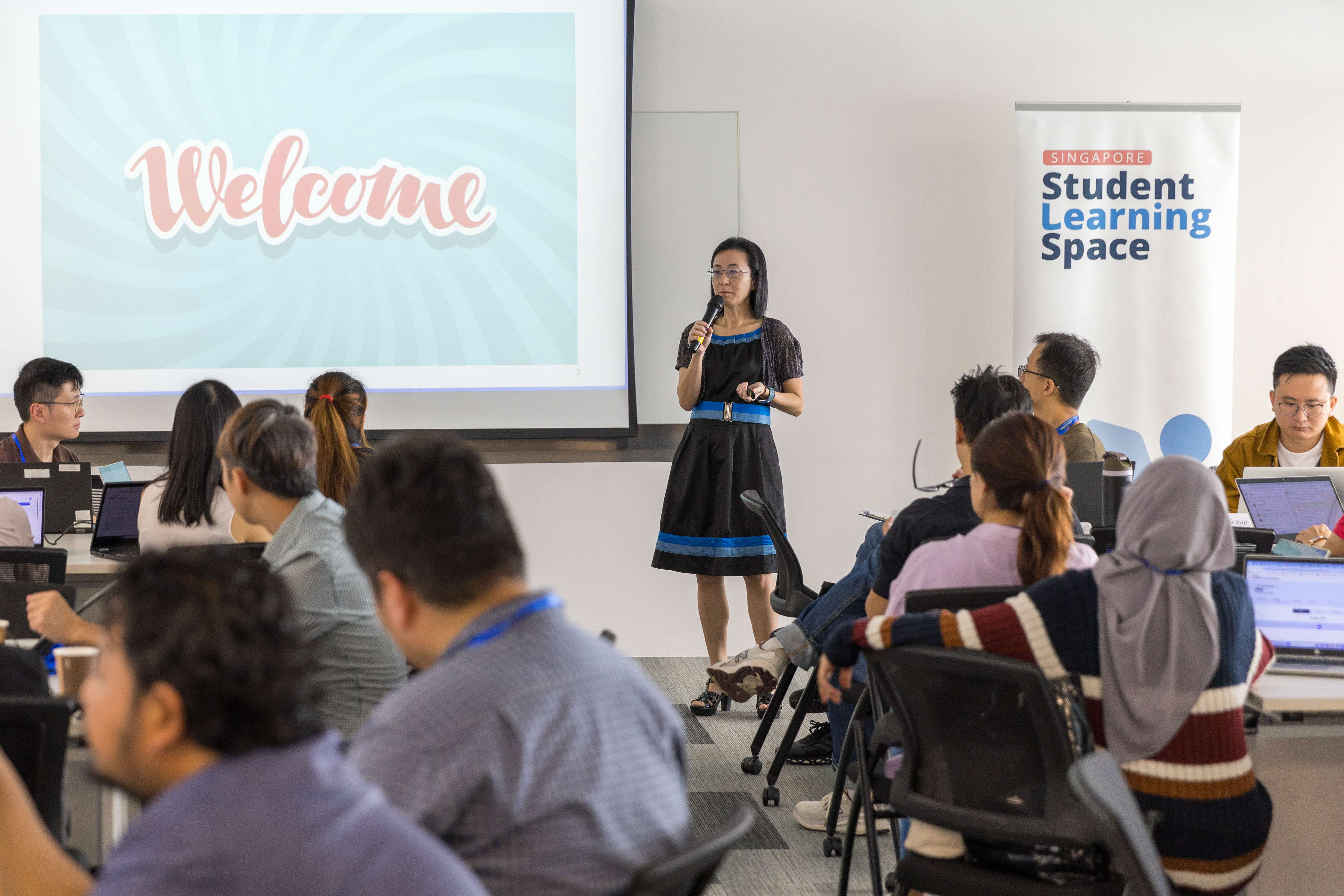 A woman holds a microphone presenting before a seated audience with a Welcome slide and Singapore Student Learning Space banner.