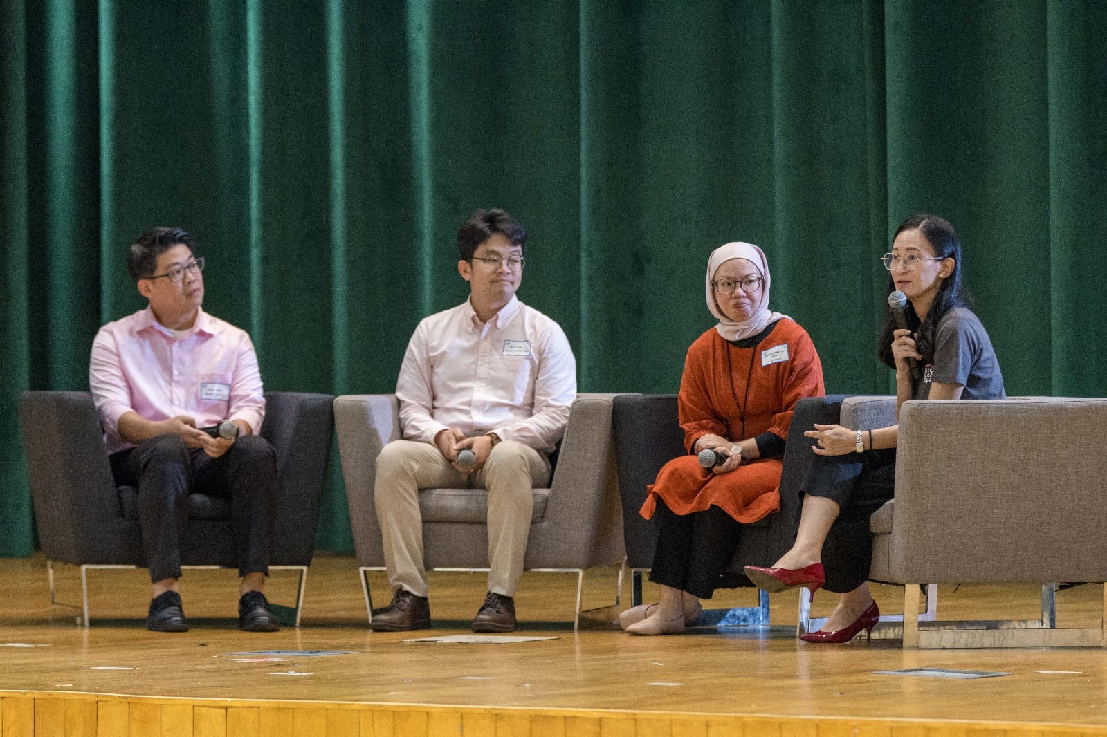 Four panelists sit on gray chairs on a wooden stage before a green curtain, each holding a microphone.