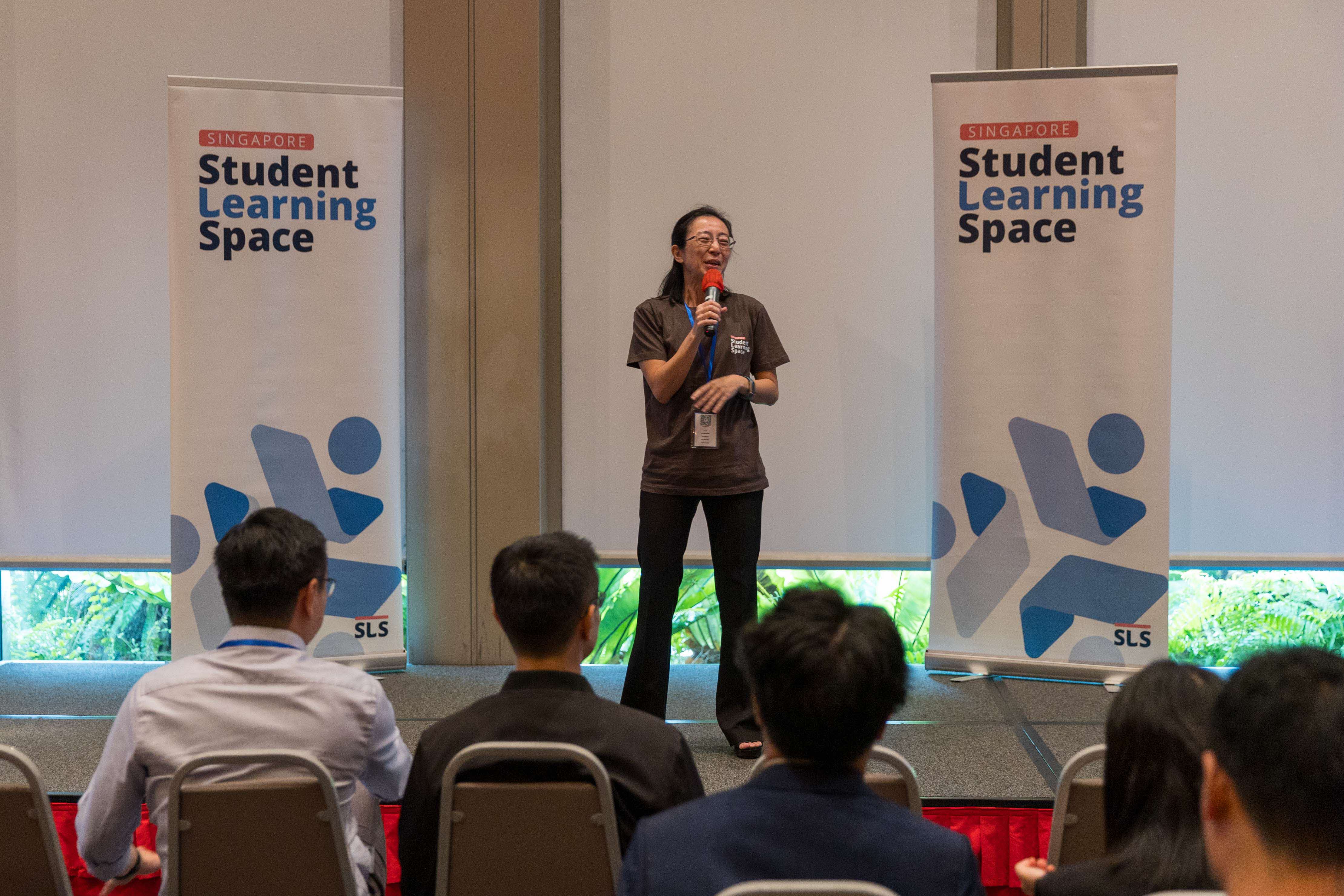 A woman speaks into a microphone on stage between two Singapore Student Learning Space banners before a seated audience.