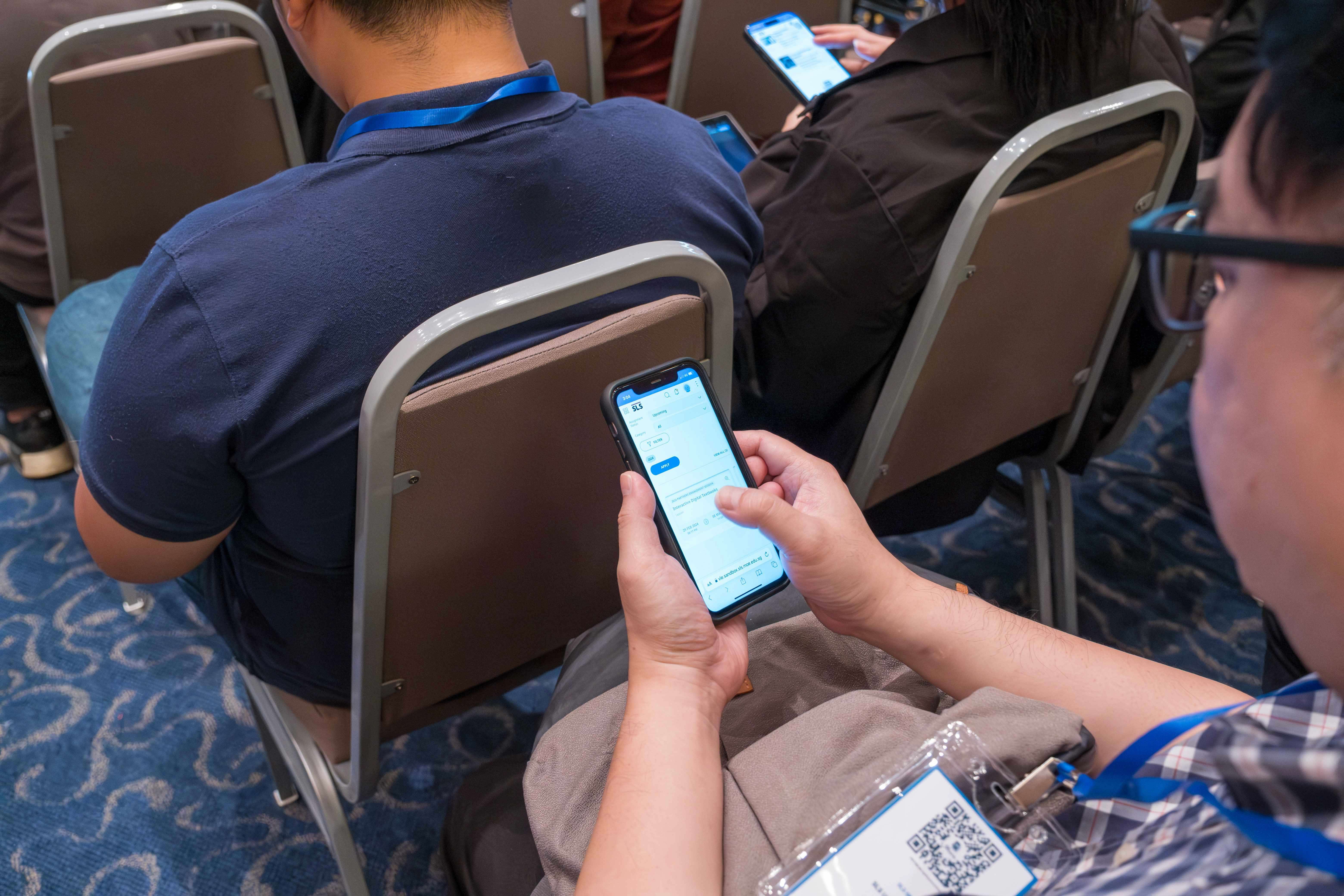 Person wearing a conference badge scrolling on a smartphone while seated in a row of chairs at an event.