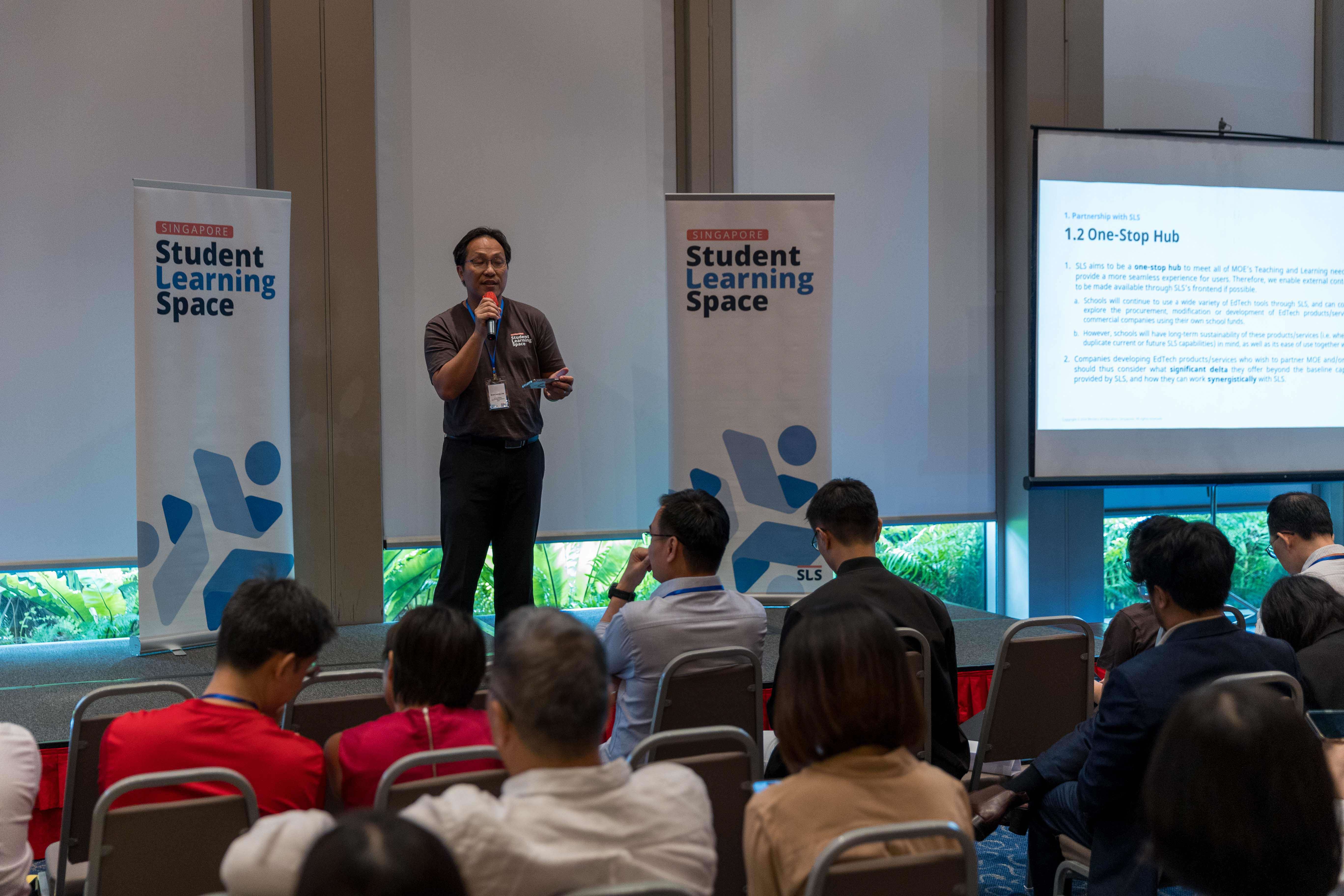 A man speaks into a microphone at a Singapore Student Learning Space event with an audience seated before him.
