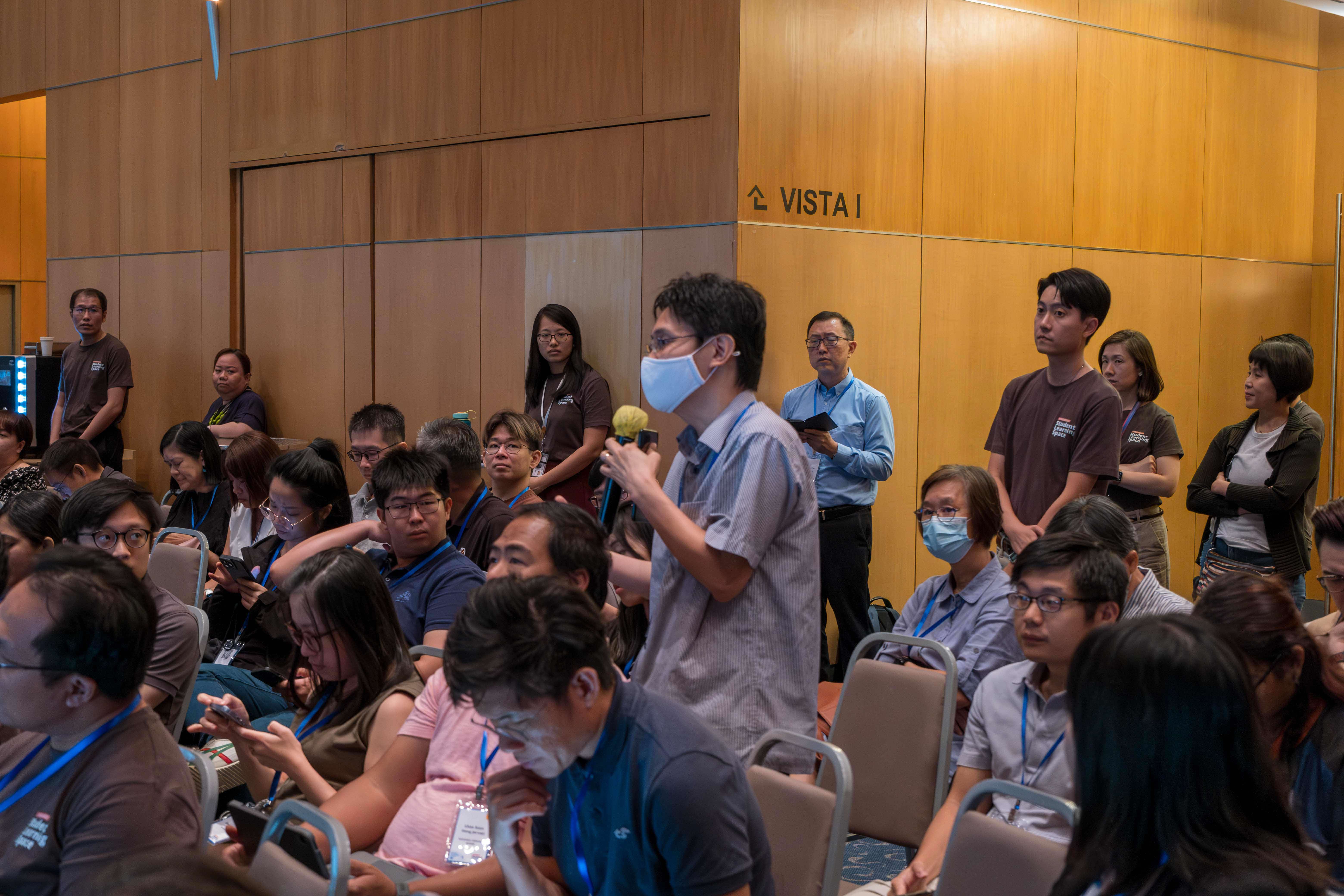 A masked man holds a microphone while standing among a seated crowd at a conference room labeled Vista I.