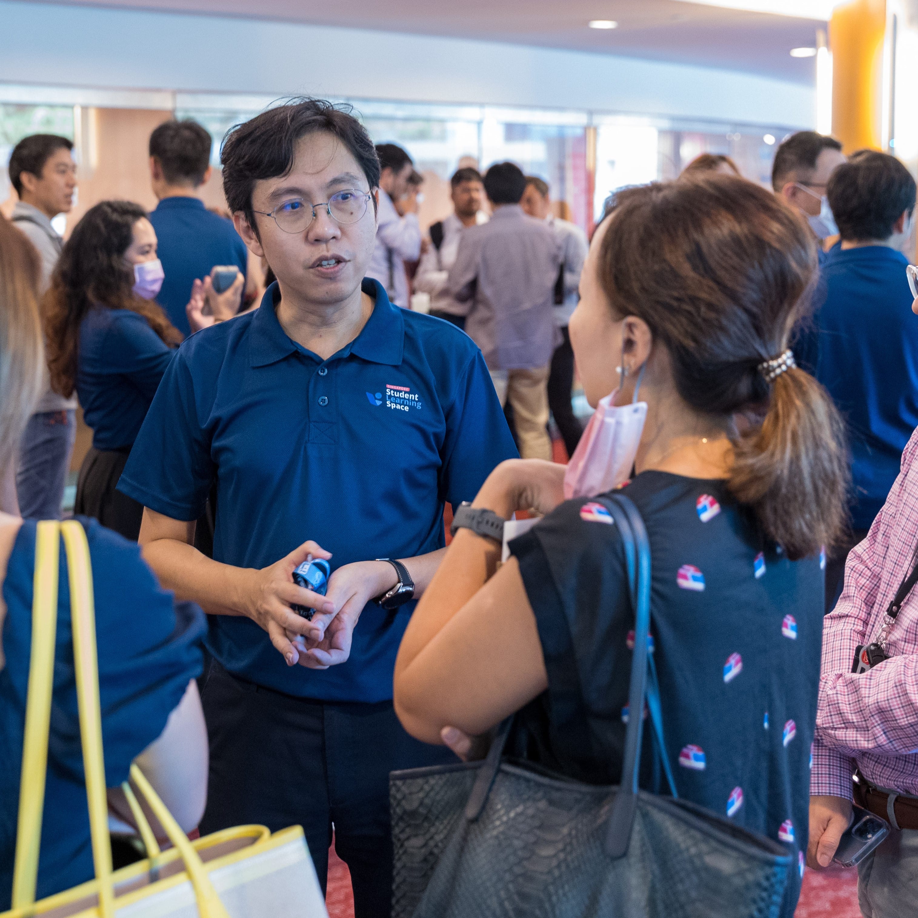 A man in a blue Student Learning Space polo shirt speaks with a woman at a busy indoor networking event.