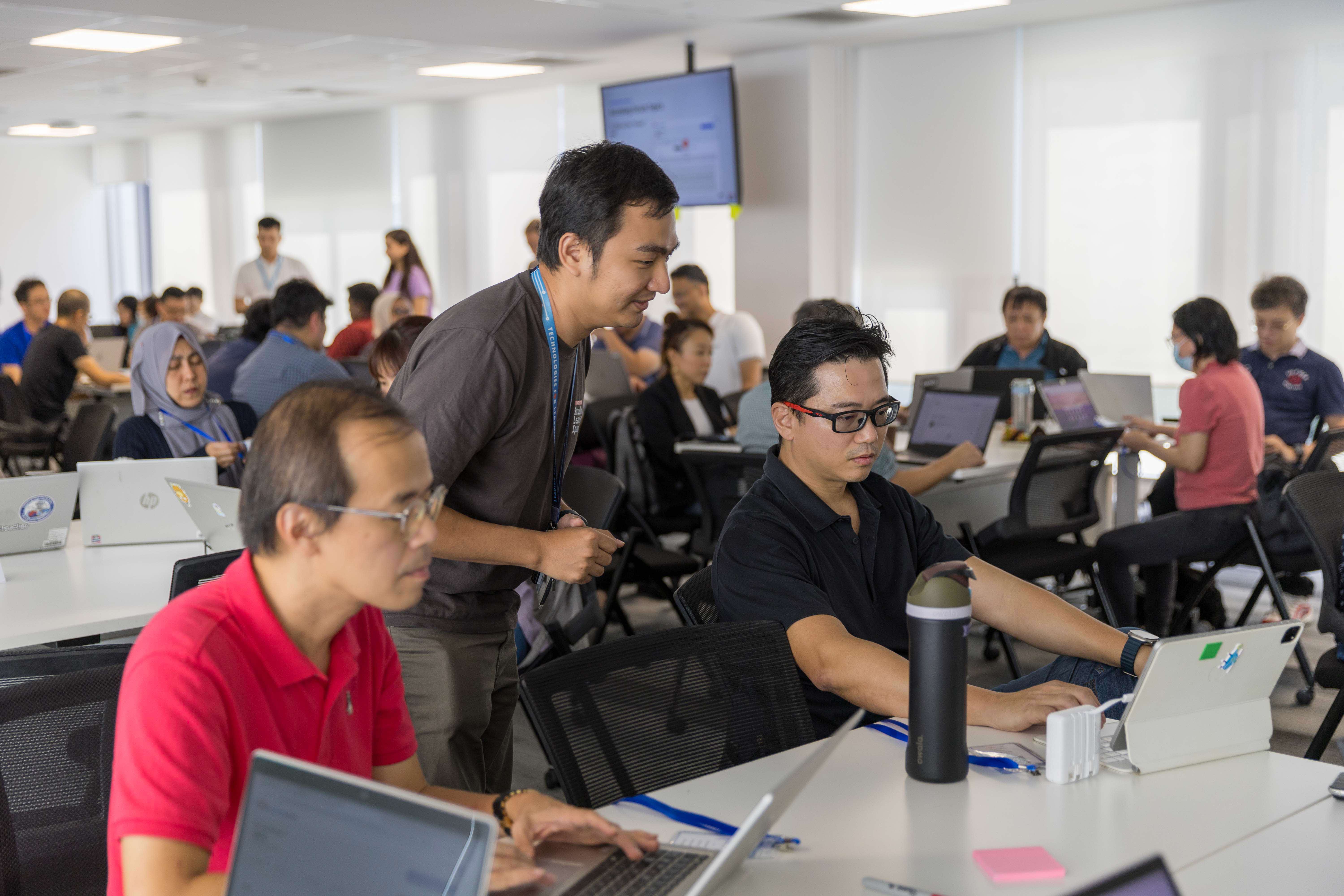 A busy workshop room with many people seated at tables working on laptops while a standing man with a lanyard assists others.
