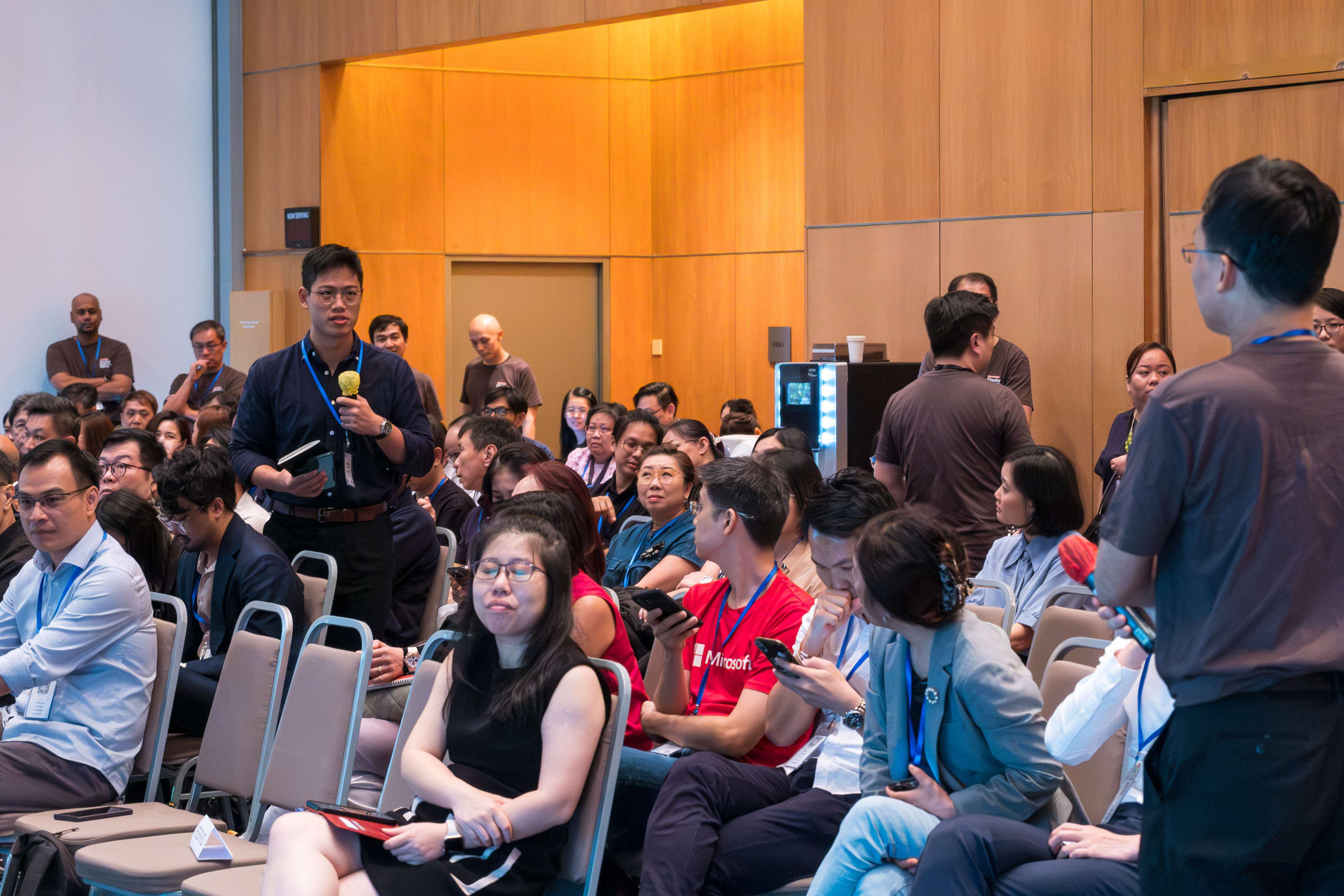 A man stands holding a microphone at a crowded indoor conference, with many seated attendees wearing lanyards around them.
