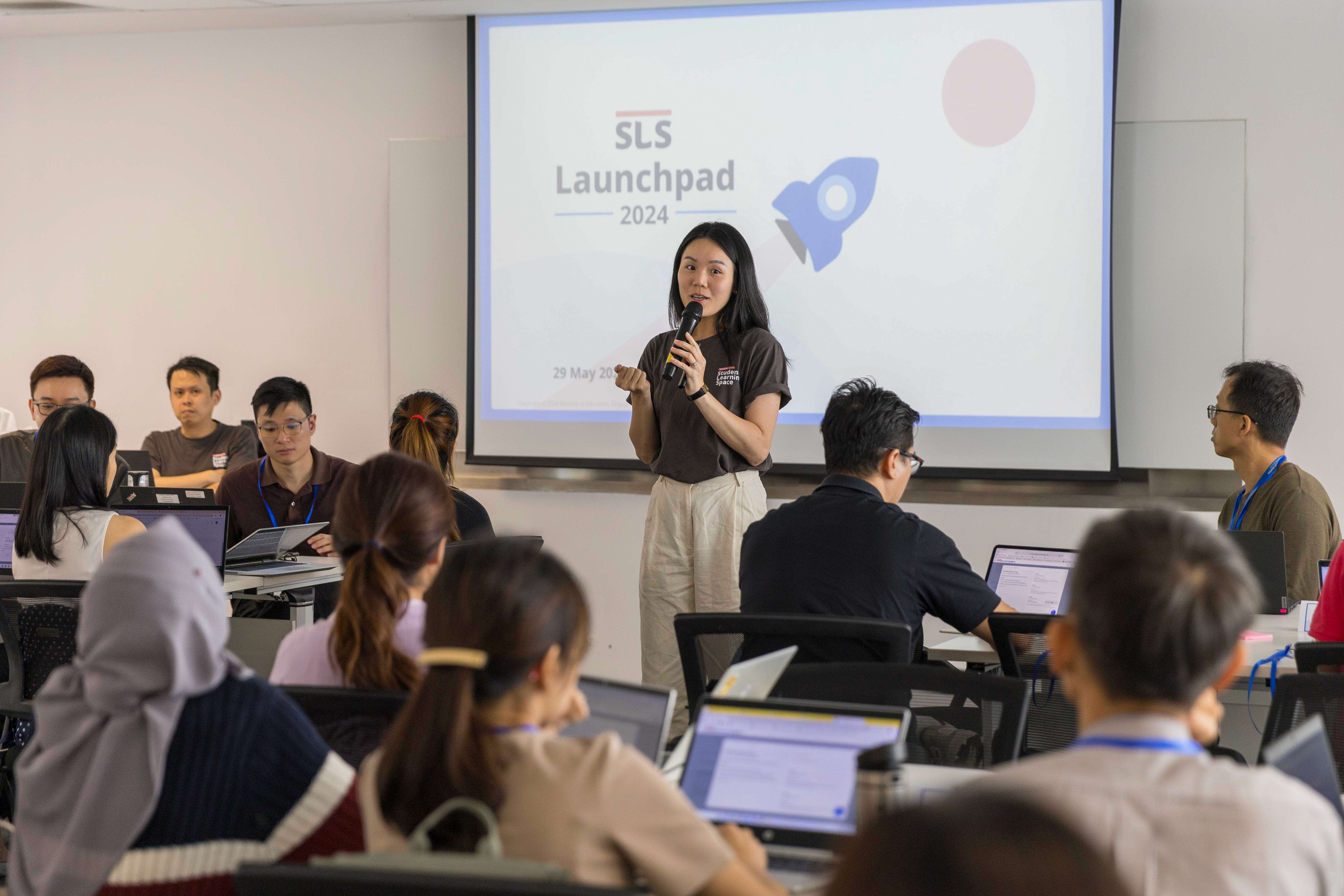 A woman speaks into a microphone before a projected slide reading "SLS Launchpad 2024" while attendees with laptops listen.