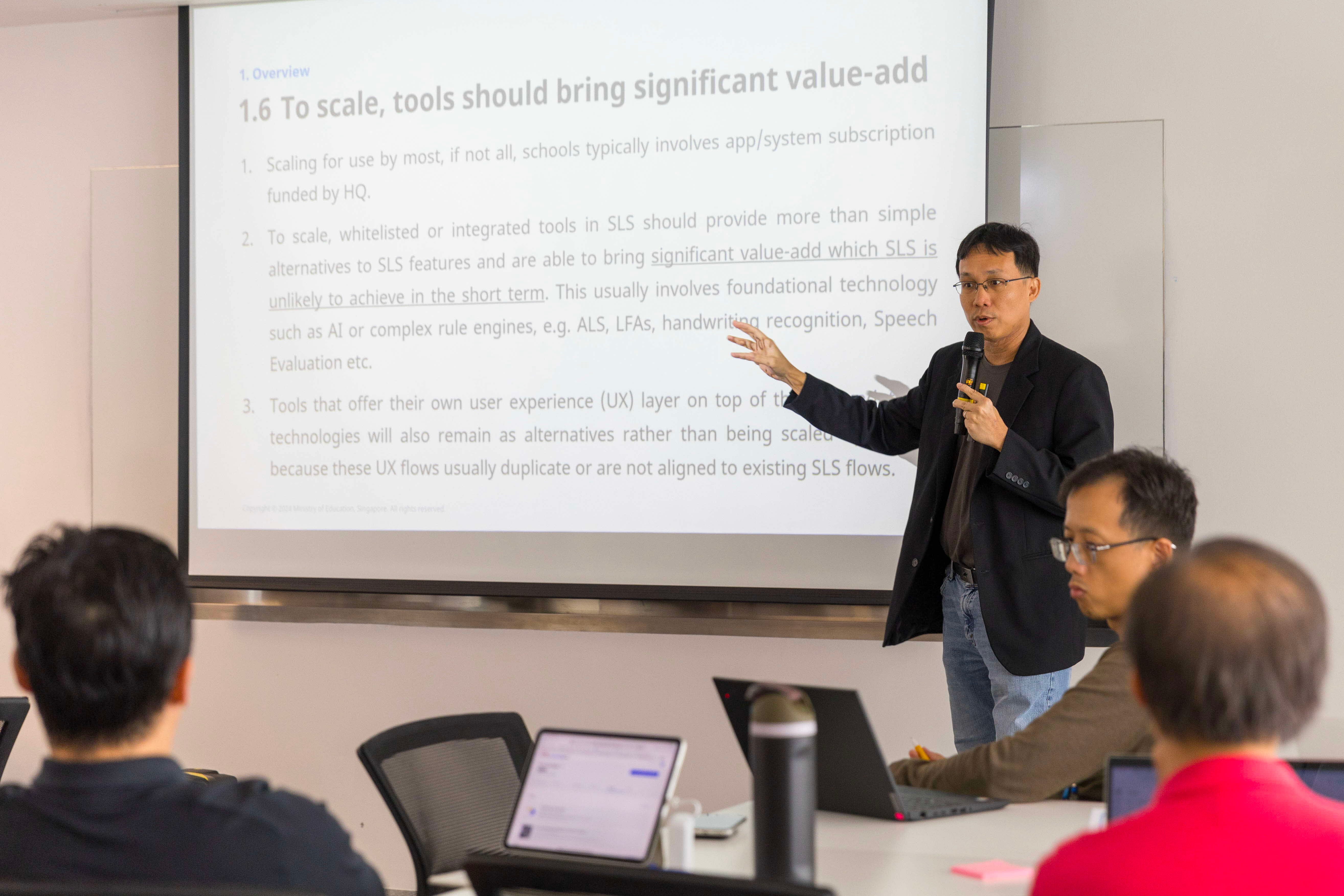 A man in a black blazer holds a microphone and gestures toward a projected slide about scaling tools in a meeting room.