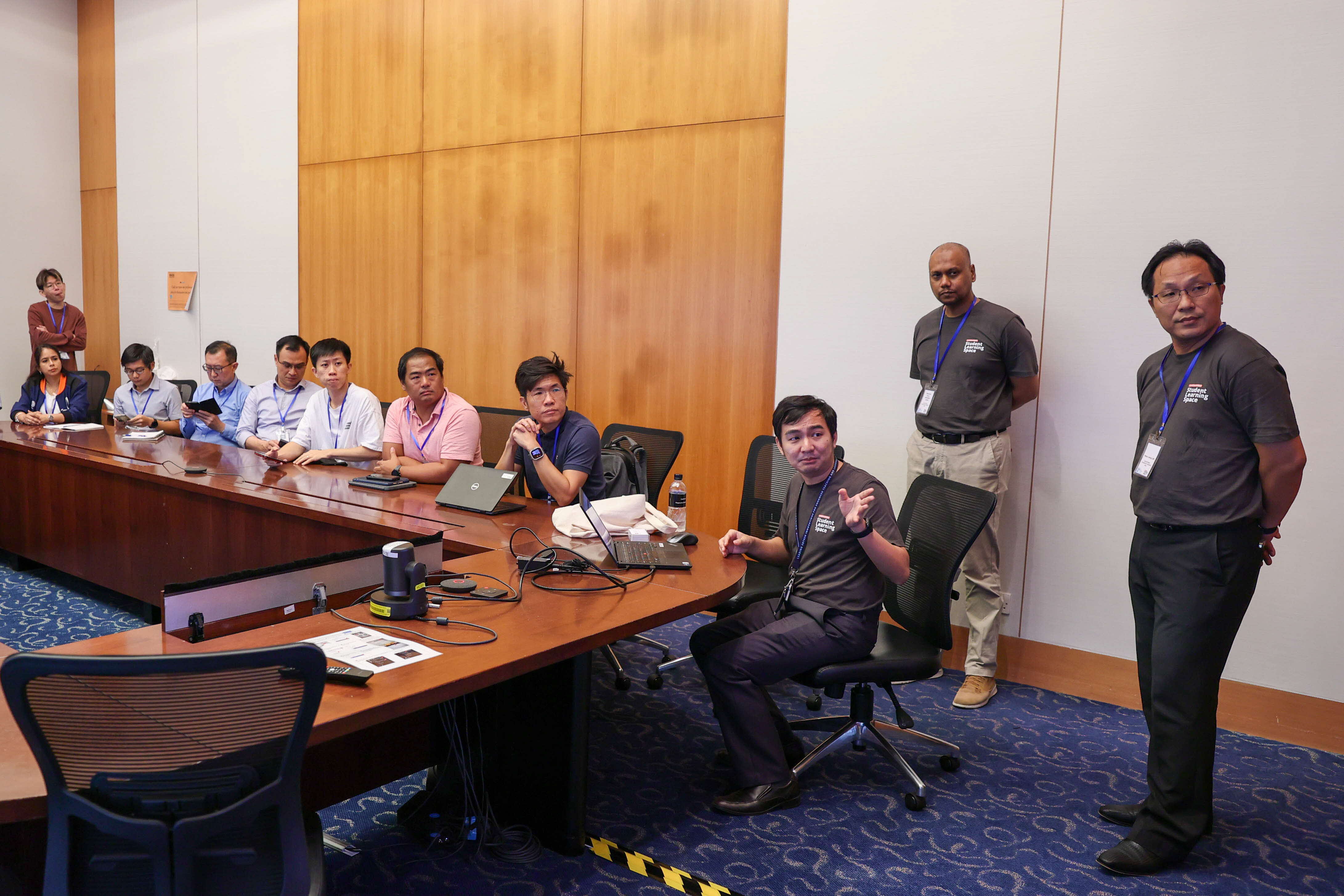 A group of people in a conference room around a wooden table with laptops, some seated and two men standing near the wall.