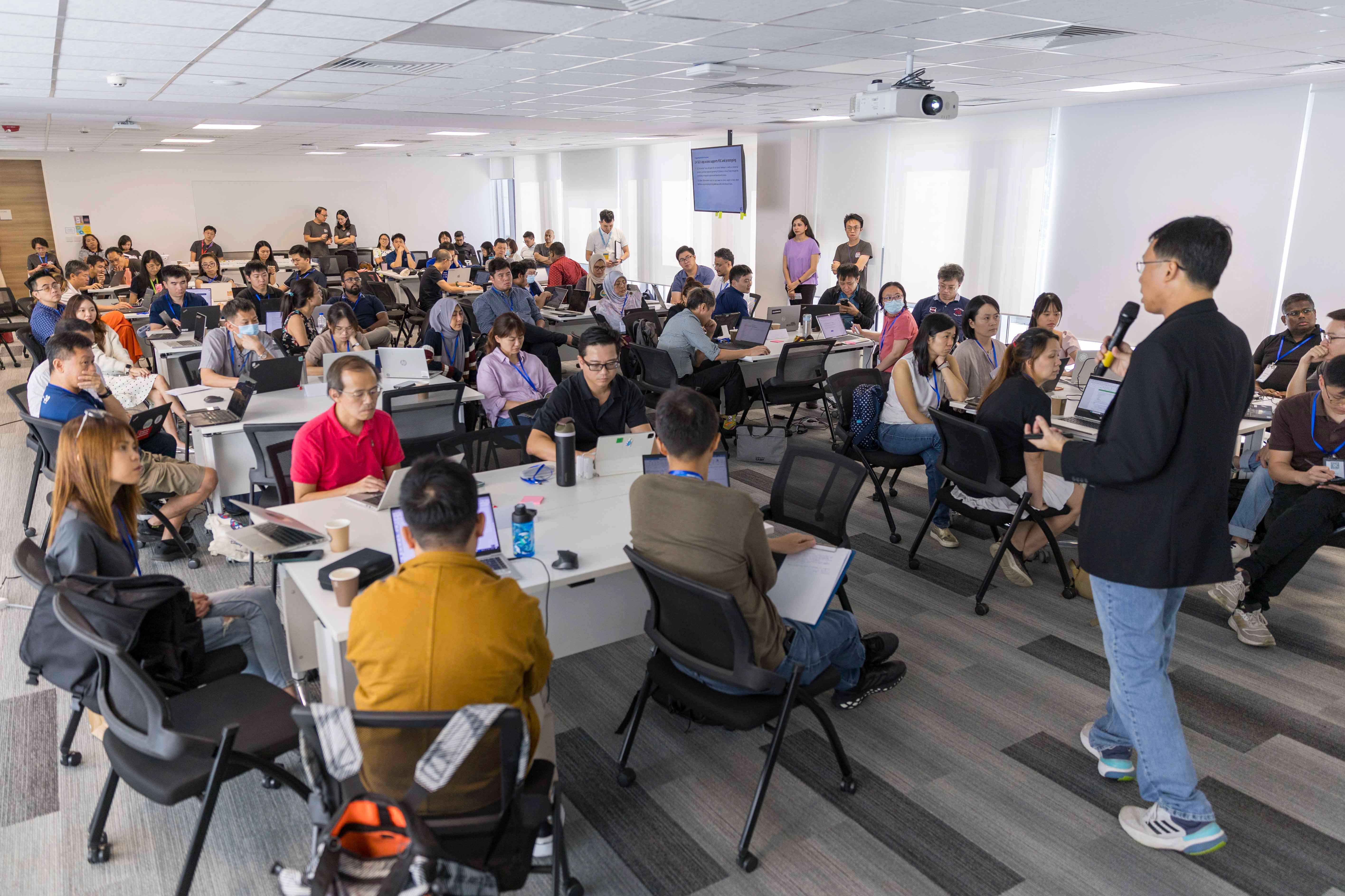 A man holding a microphone addresses a large group of people seated at tables with laptops in a bright conference room.