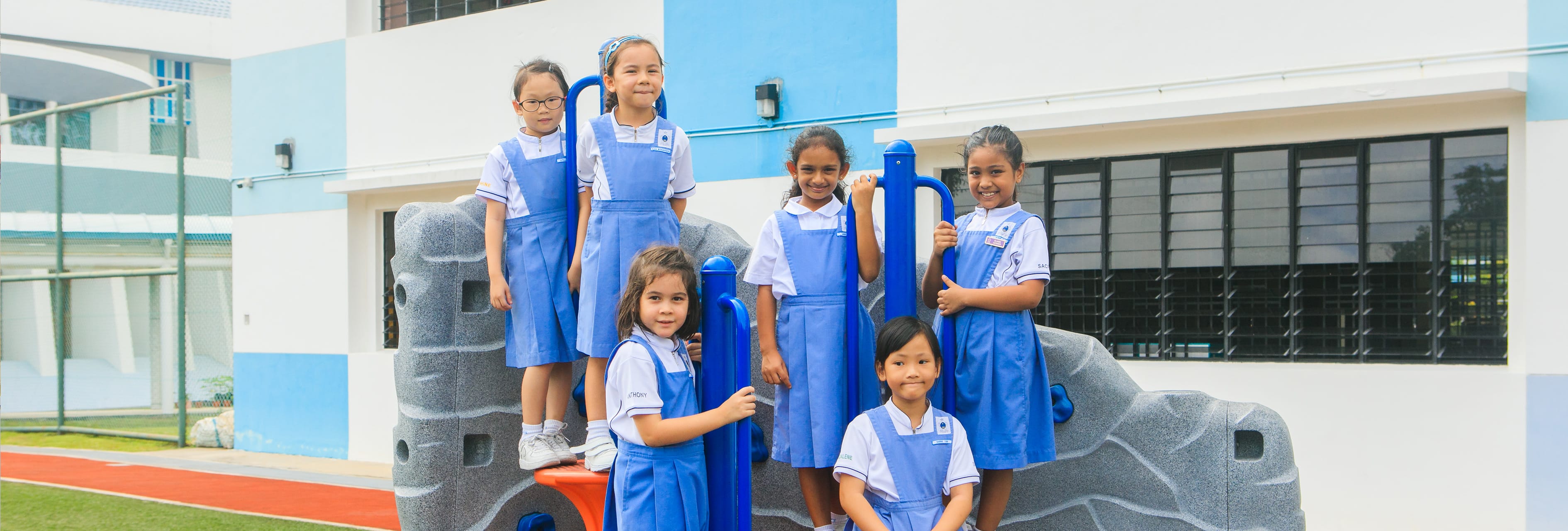 Six young girls in blue and white school uniforms smile on a playground climbing structure outside a school building.