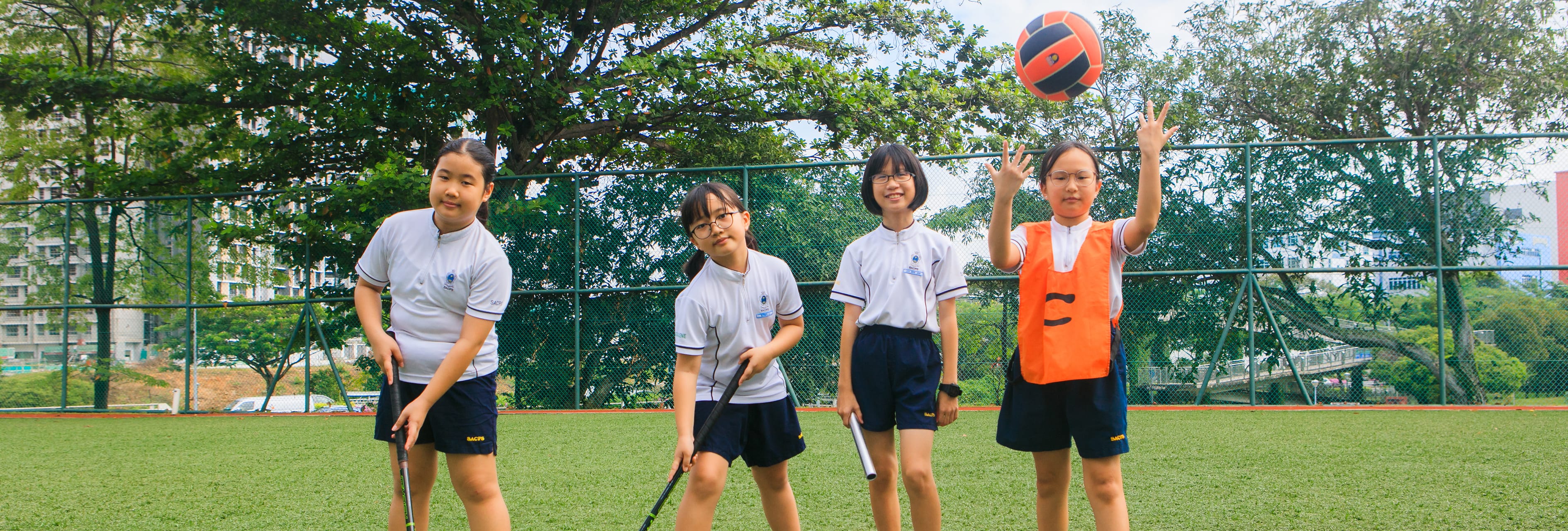 Four students on a sports field; one in orange bib tosses a ball while two hold hockey sticks.