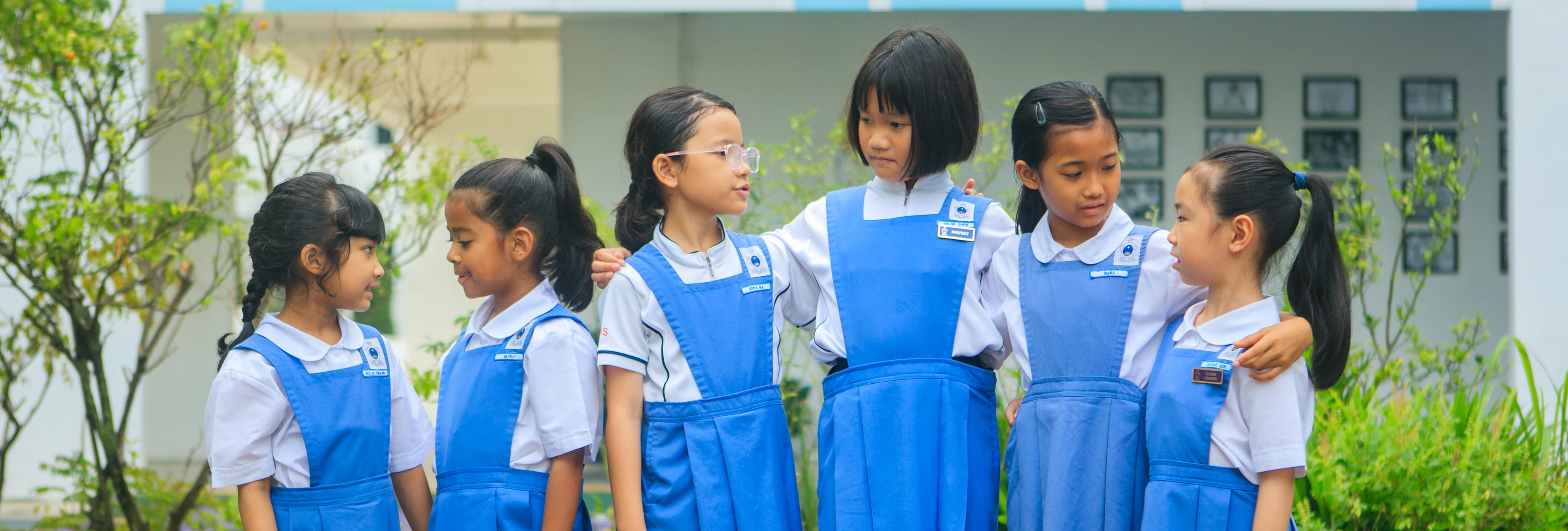Six young girls in blue and white school uniforms stand together outdoors near a building, some with arms around each other.