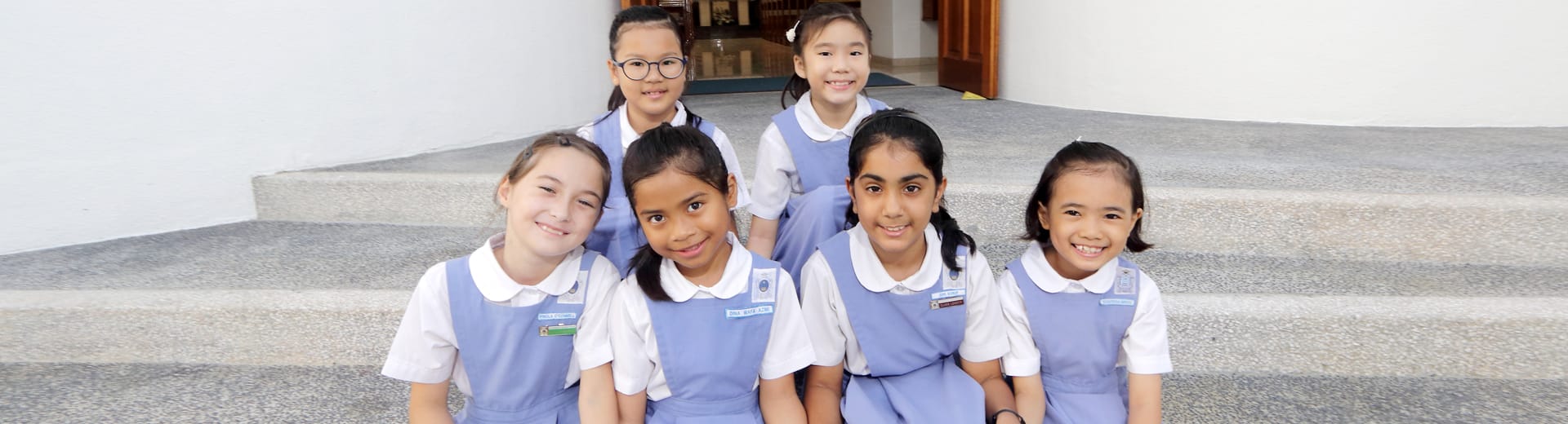 Six young girls in blue and white school uniforms smile while sitting on stone steps.