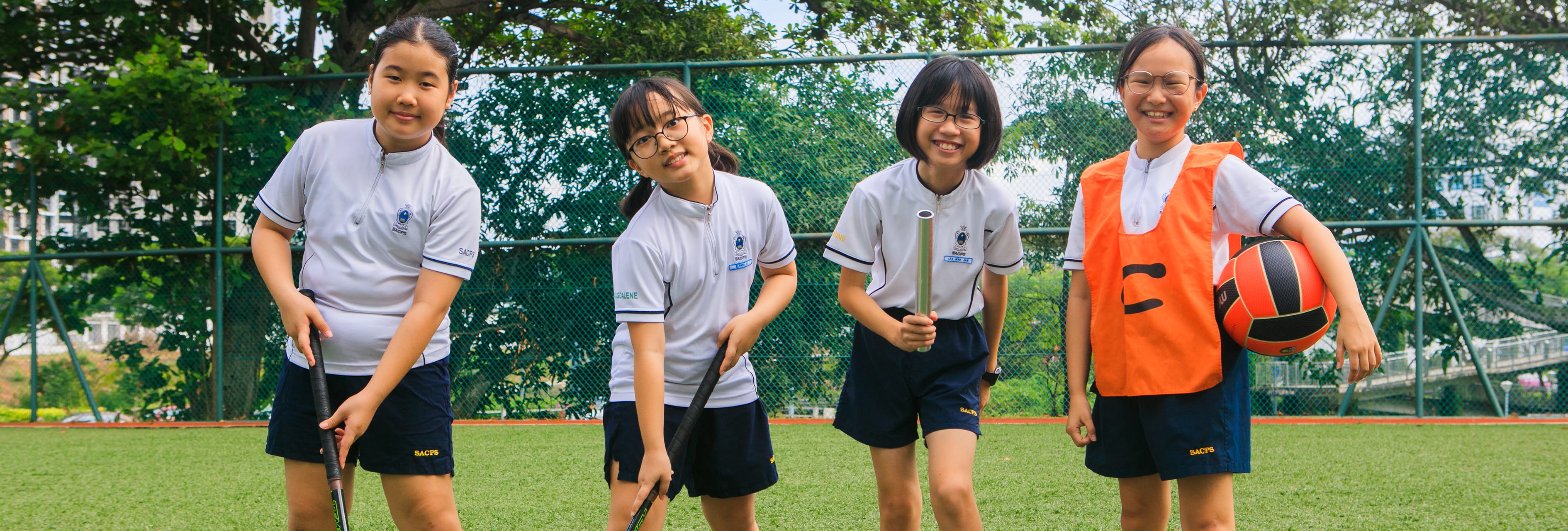 Four schoolgirls on a field; three hold hockey sticks and one in an orange bib holds a basketball.