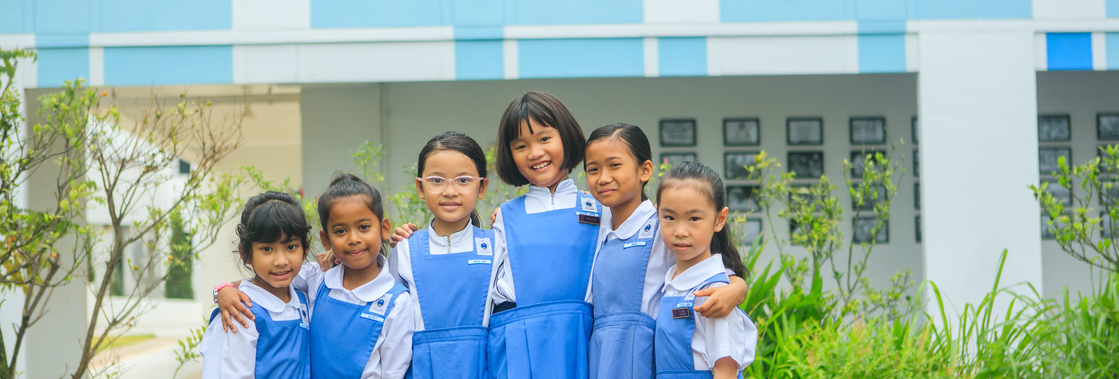 Six young girls in blue and white school uniforms smile together outdoors in front of a blue and white building.