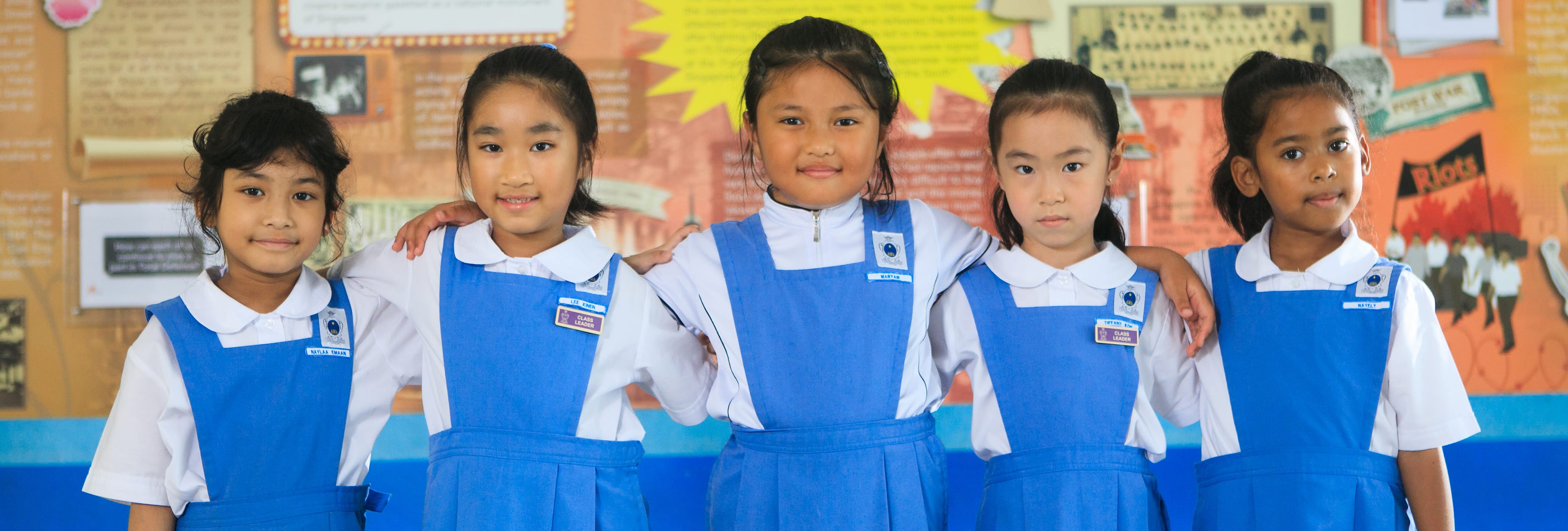 Five young girls in blue and white school uniforms stand together smiling in front of a colorful classroom display.