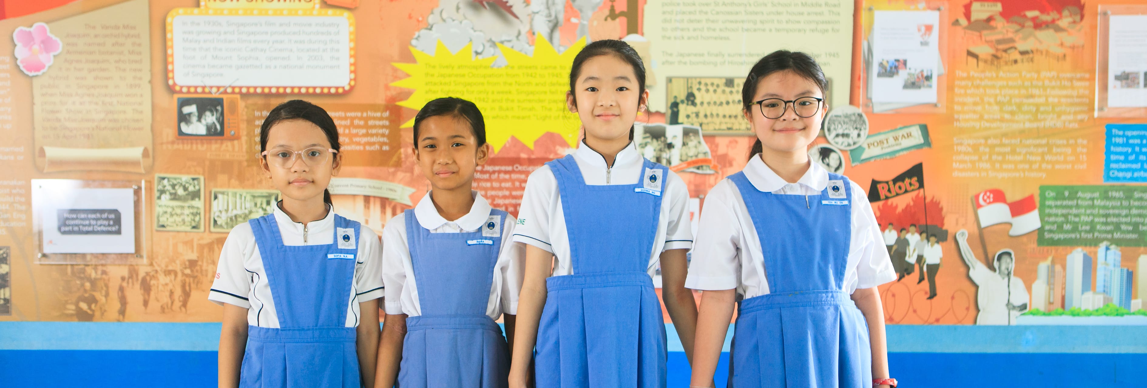 Four young girls in blue and white school uniforms smile in front of a colorful history display board.