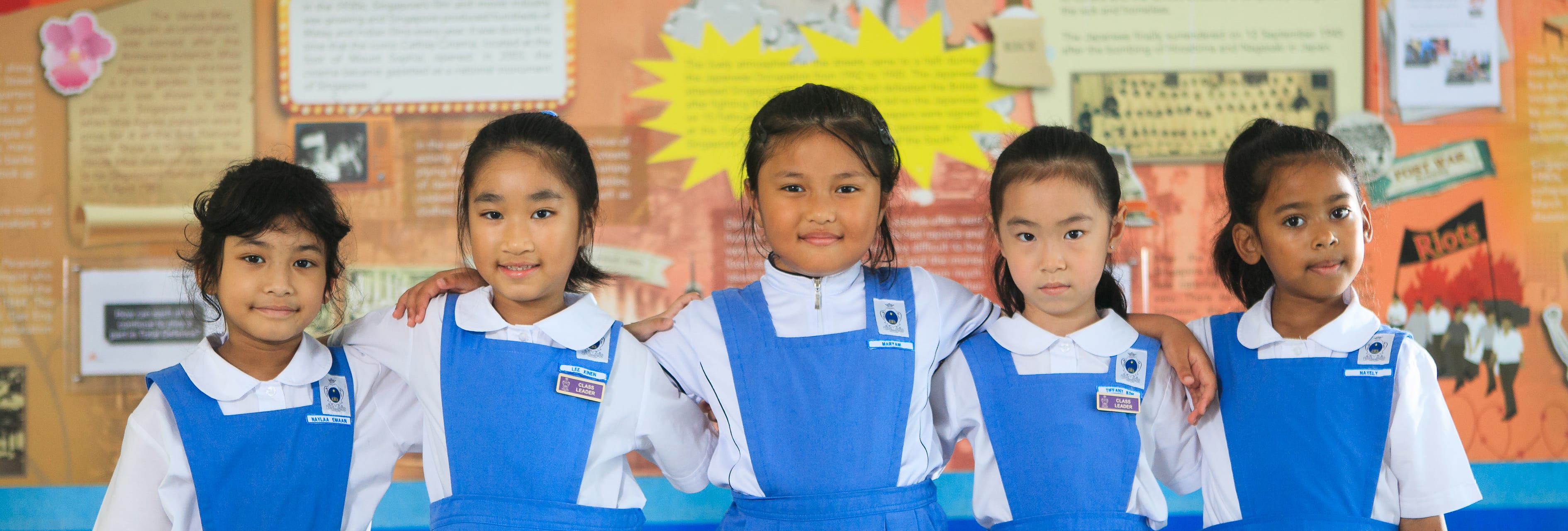Five young girls in blue and white school uniforms stand together smiling in front of a colorful classroom display board.