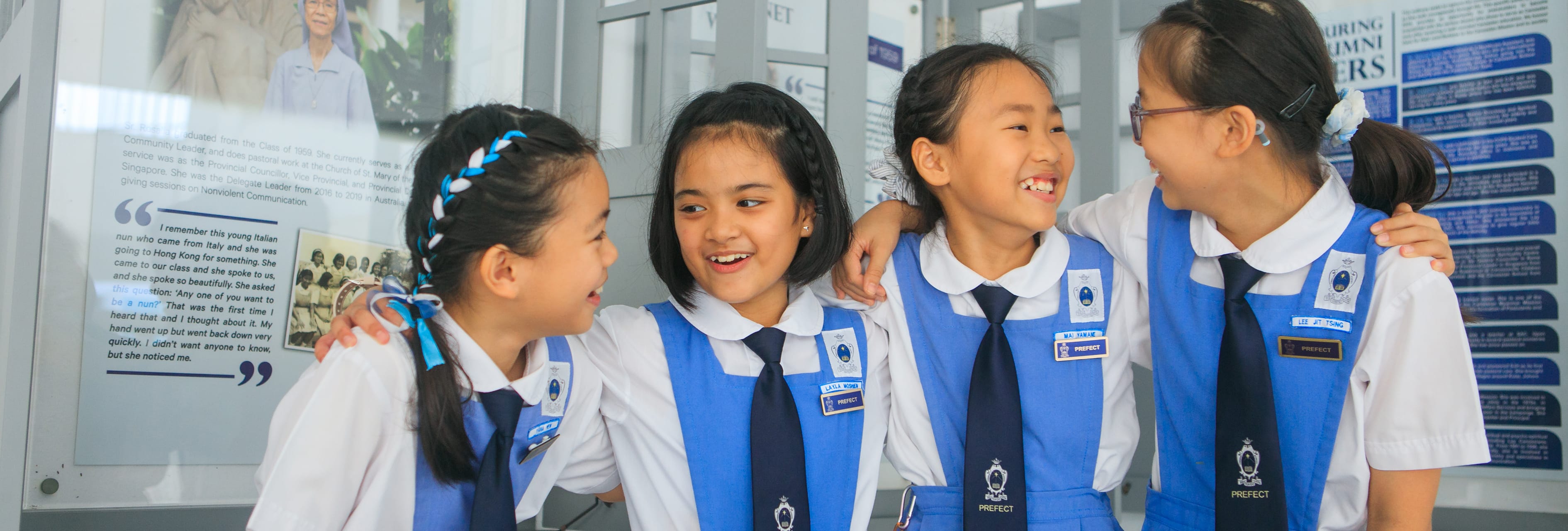 Four smiling schoolgirls in blue and white uniforms with prefect badges laugh together near a display board.