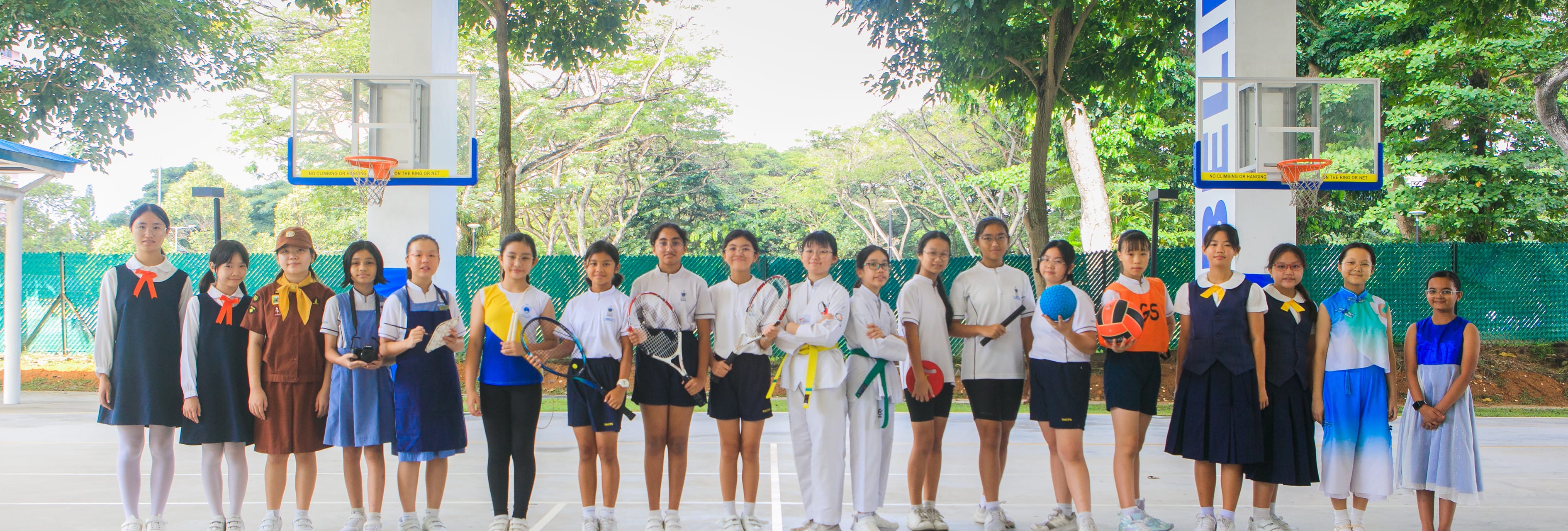 Line of schoolgirls in uniforms and sports gear holding rackets and balls on an outdoor basketball court.