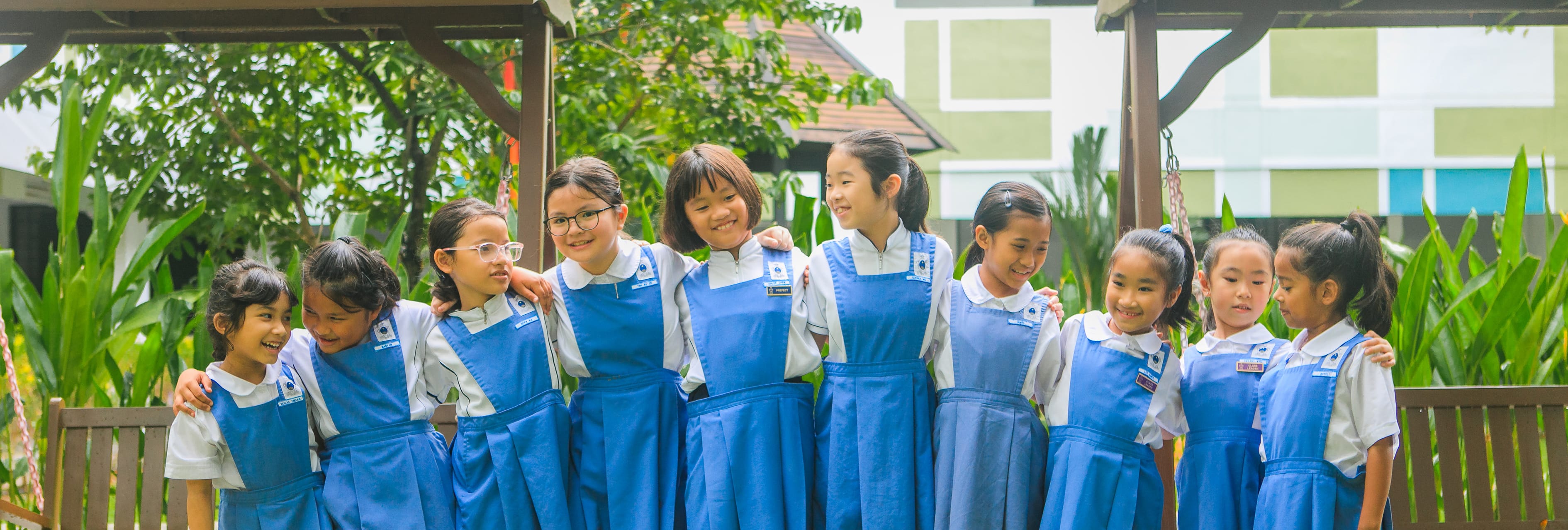Nine schoolgirls in blue uniforms stand arm in arm outdoors, smiling and chatting under a wooden frame.